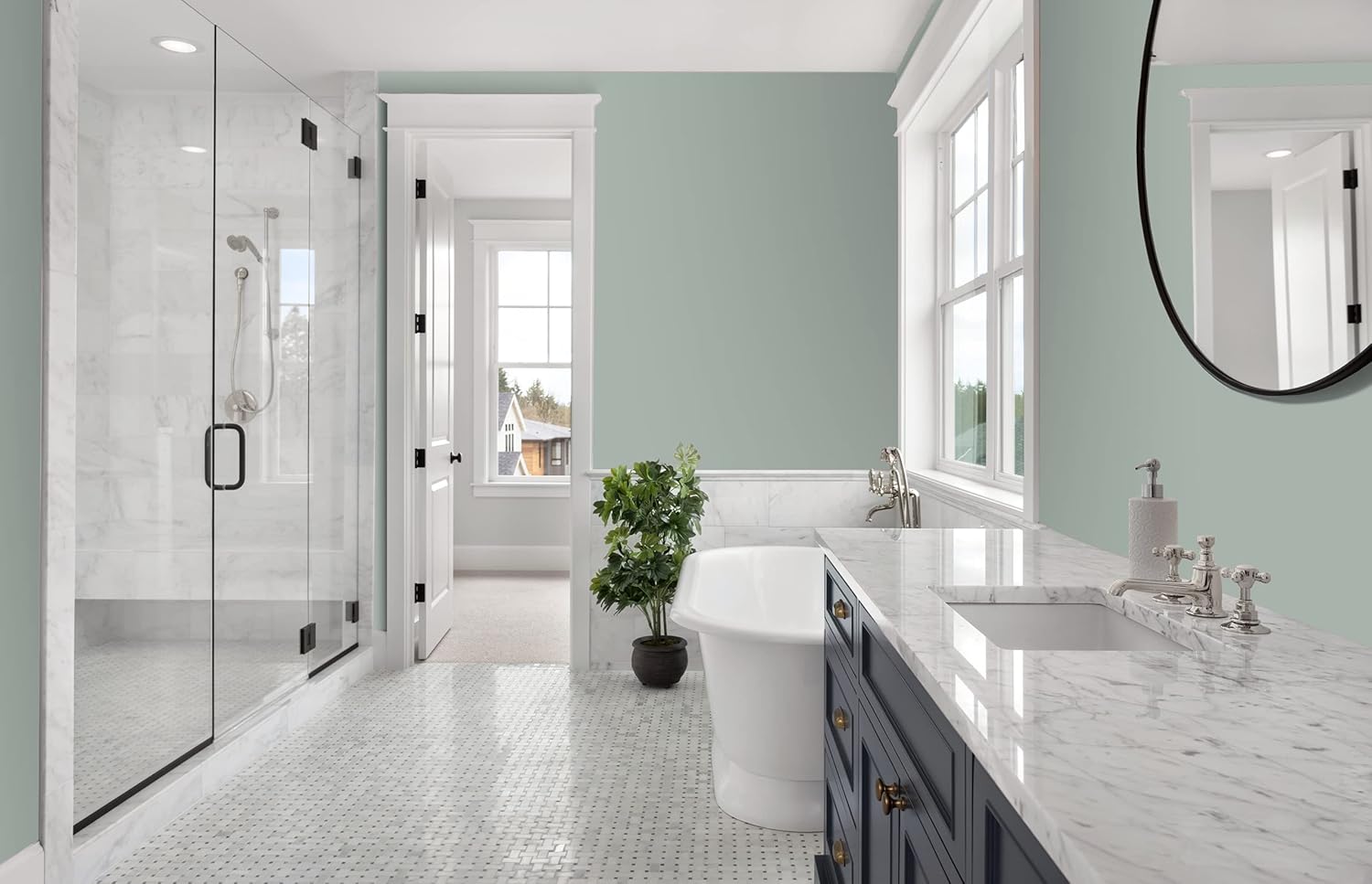 Modern bathroom with light green walls in a thick satin finish paint, a glass shower, freestanding white bathtub, marble countertops, blue vanity, round mirror, mosaic tile floor, large windows, and a potted plant by the tub.