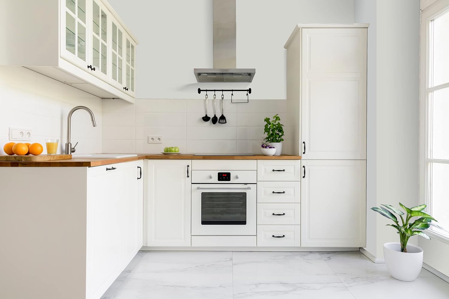 A bright, modern kitchen featuring white cabinets, a wooden countertop, and a stainless steel oven. A potted plant and a bowl of oranges add a touch of greenery and color to the space. The room has large windows, allowing natural light to fill the area.