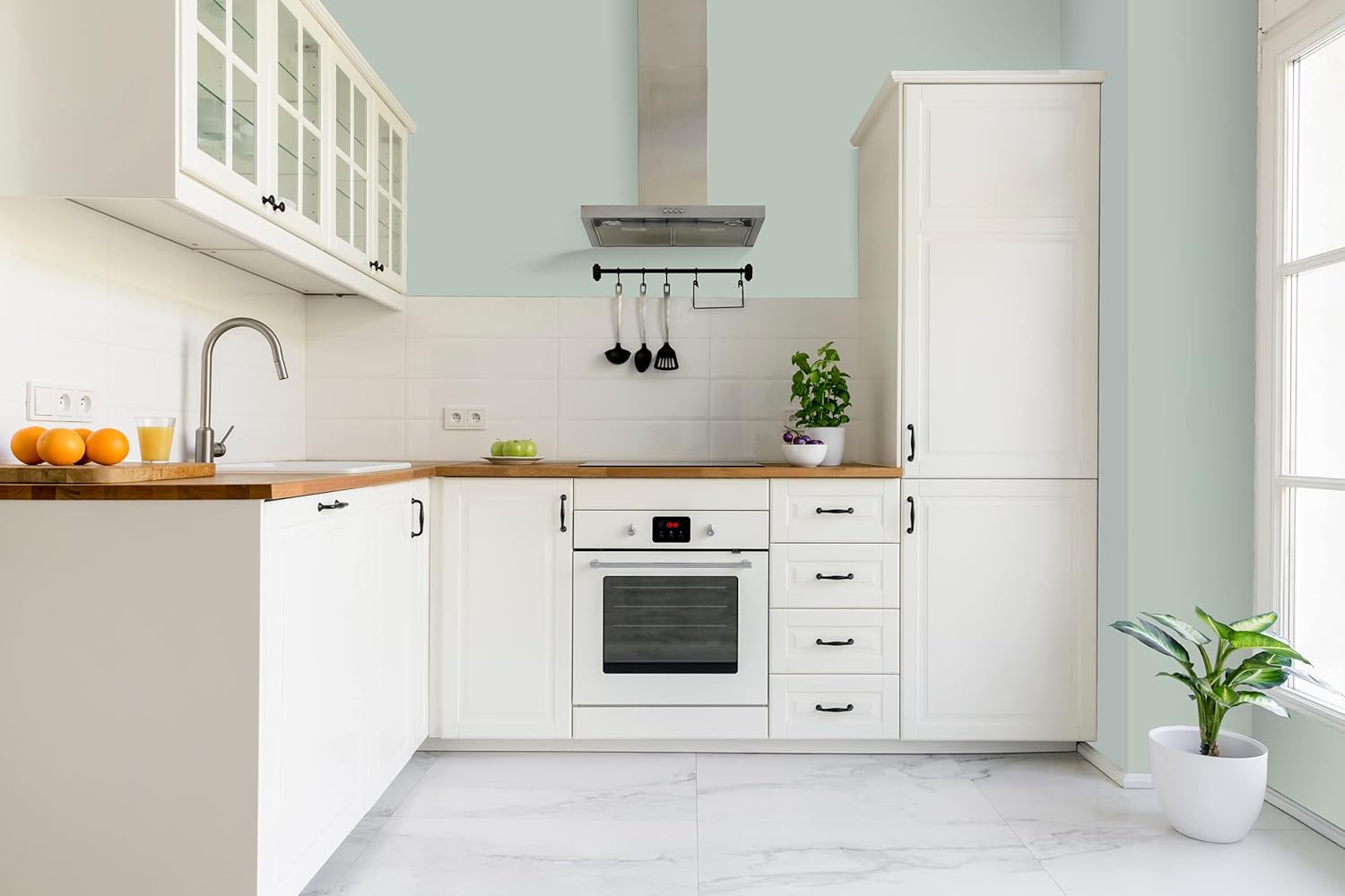 Modern kitchen with white cabinets finished in premium paint, wooden countertops, a built-in oven, a sink, and a range hood. Kitchen utensils hang on the wall and potted plants add freshness to the space.