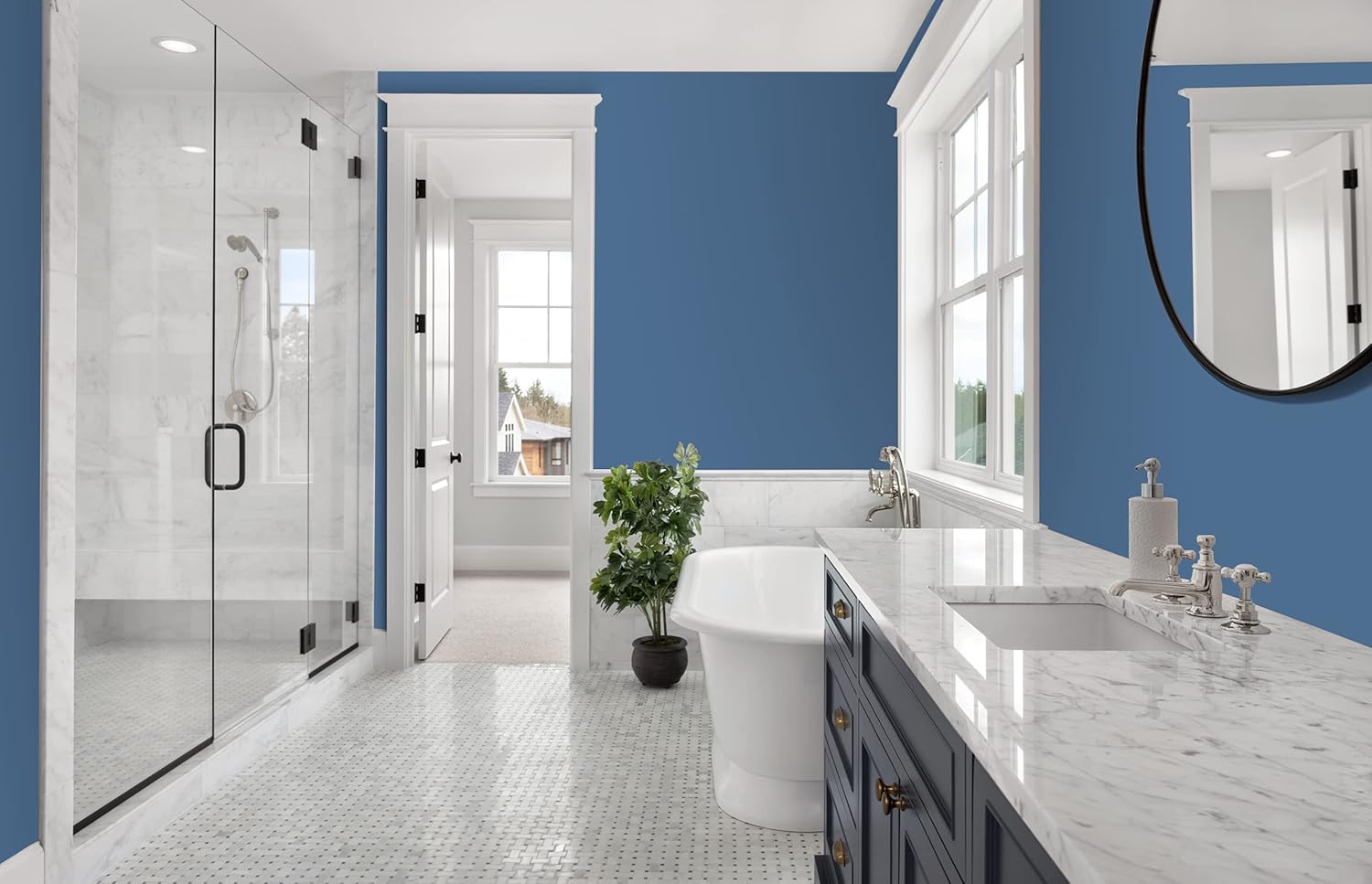 Modern bathroom with blue walls coated in thick satin finish paint, large windows, marble countertop and floor, freestanding bathtub, glass shower enclosure, round mirror, and a potted plant near the tub. Bright and spacious interior.