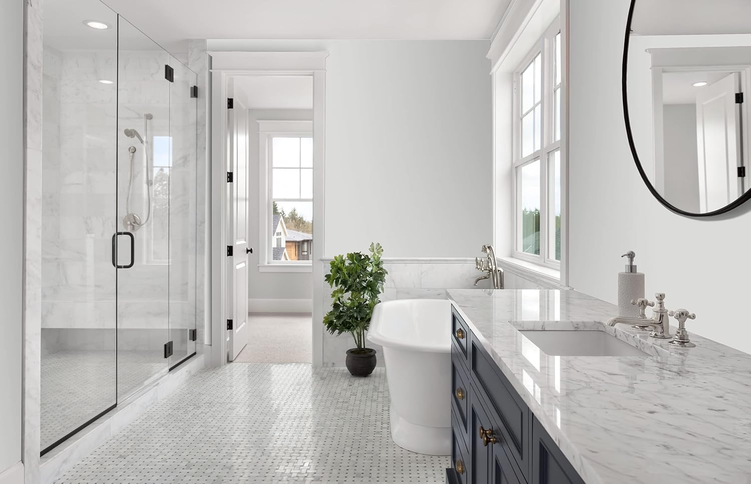 A modern bathroom featuring a walk-in glass shower, a freestanding bathtub, and a marble countertop with dual sinks. Natural light floods the space through large windows, complemented by a potted plant for a touch of greenery.