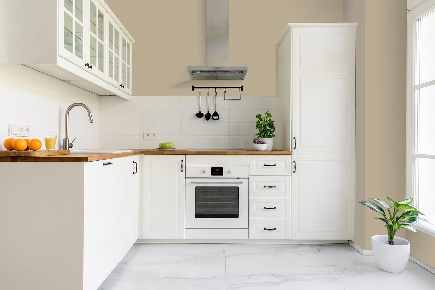 Modern kitchen with white cabinets finished in premium paint, wooden countertops, stainless steel oven and range hood, potted plants, utensils hanging on hooks, a bowl of oranges, and large window letting in natural light.