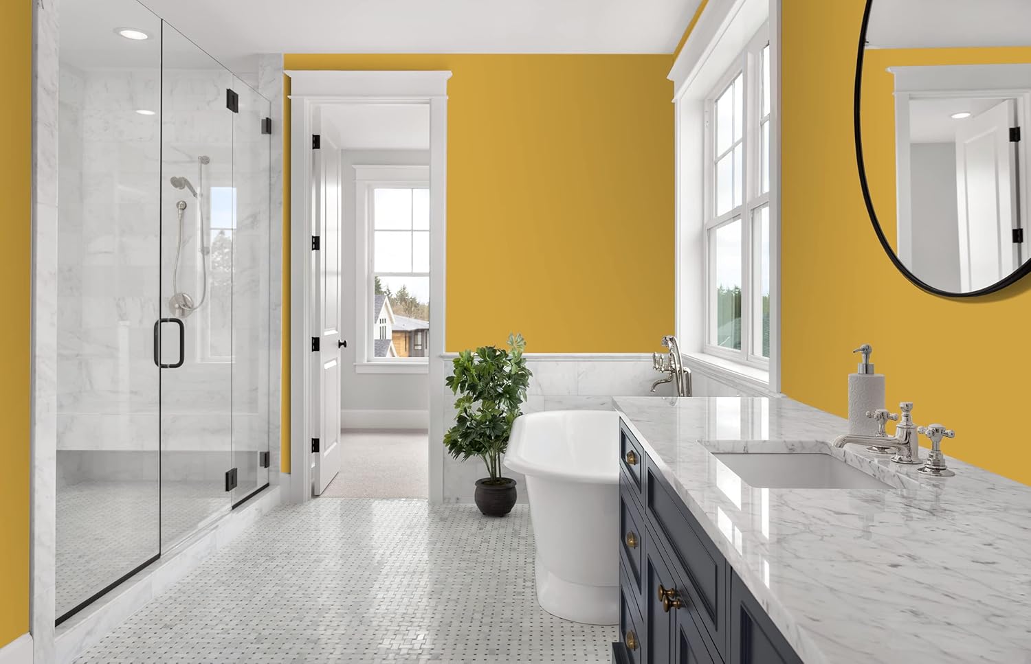 Modern bathroom with yellow walls painted in a thick satin finish paint, white trim, and marble surfaces. Features a glass shower, freestanding tub, navy blue vanity, round mirror, and large windows letting in natural light. A potted plant adds greenery.