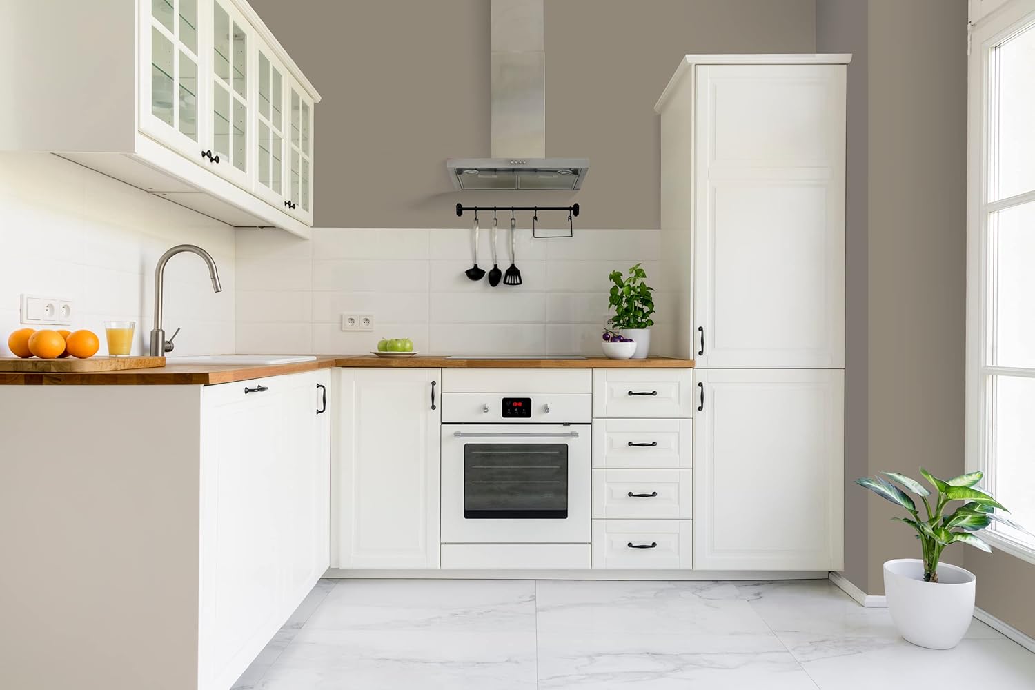 Modern kitchen with white cabinets finished in thick satin finish paint, wooden countertops, built-in oven, sink, potted plants, and utensils hanging under a range hood. Sunlight streams in from a window on the right.