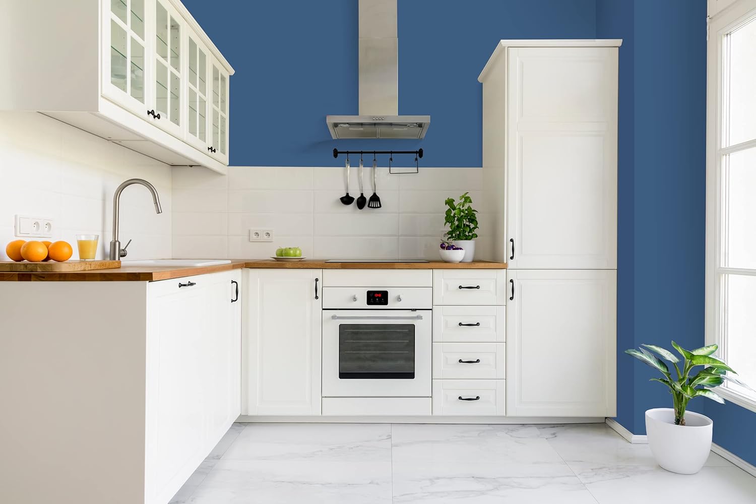 Modern kitchen with white cabinets, wooden countertops, stainless steel oven and hood, utensils hanging on a rack, potted plants, and blue accent walls in a thick satin finish paint. Sunlight enters through a window on the right.