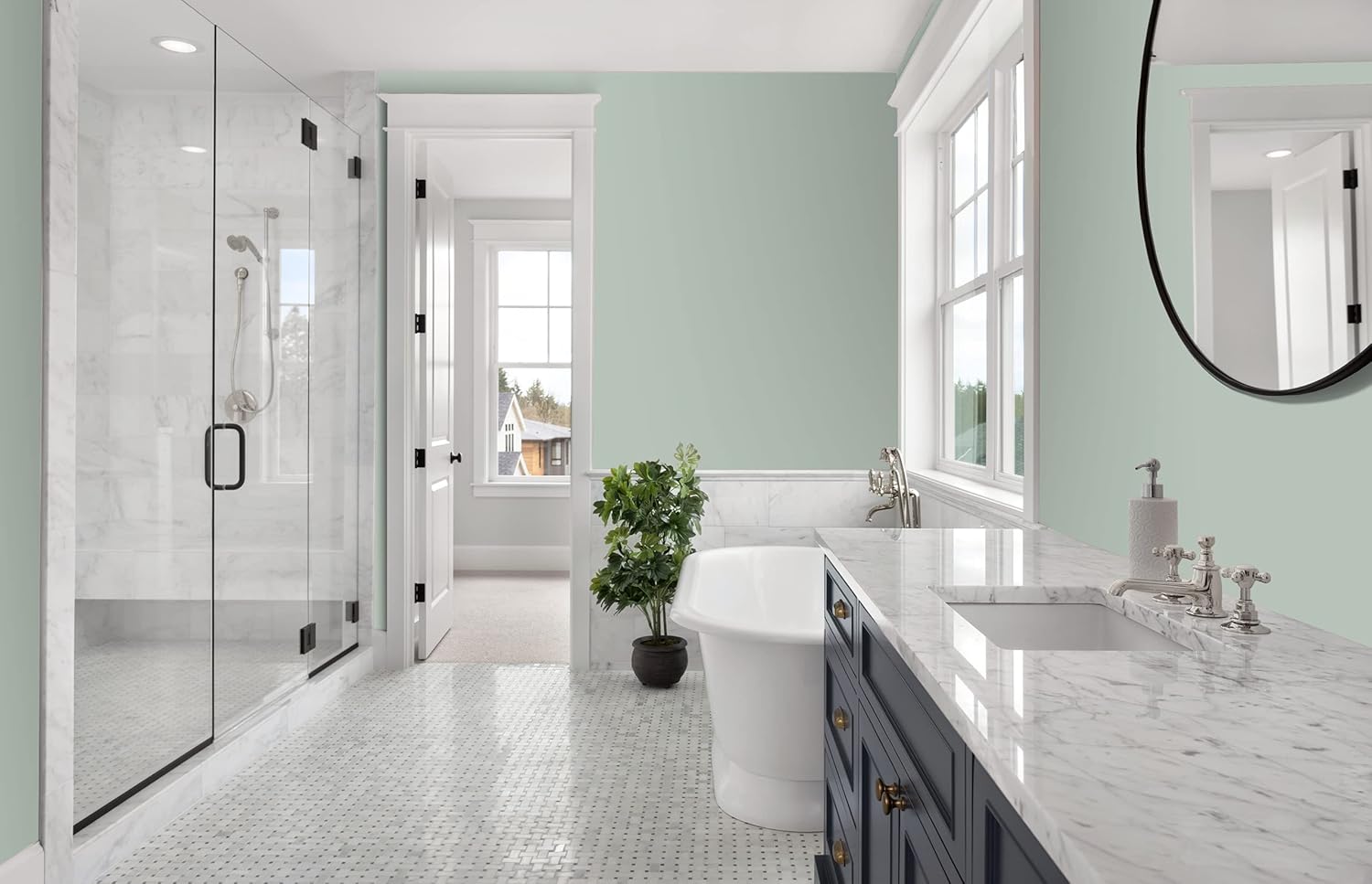 A bright, modern bathroom featuring a glass shower, a freestanding bathtub, and a marble countertop with dual sinks. Soft green walls complement large windows that allow natural light. A potted plant adds a touch of greenery.