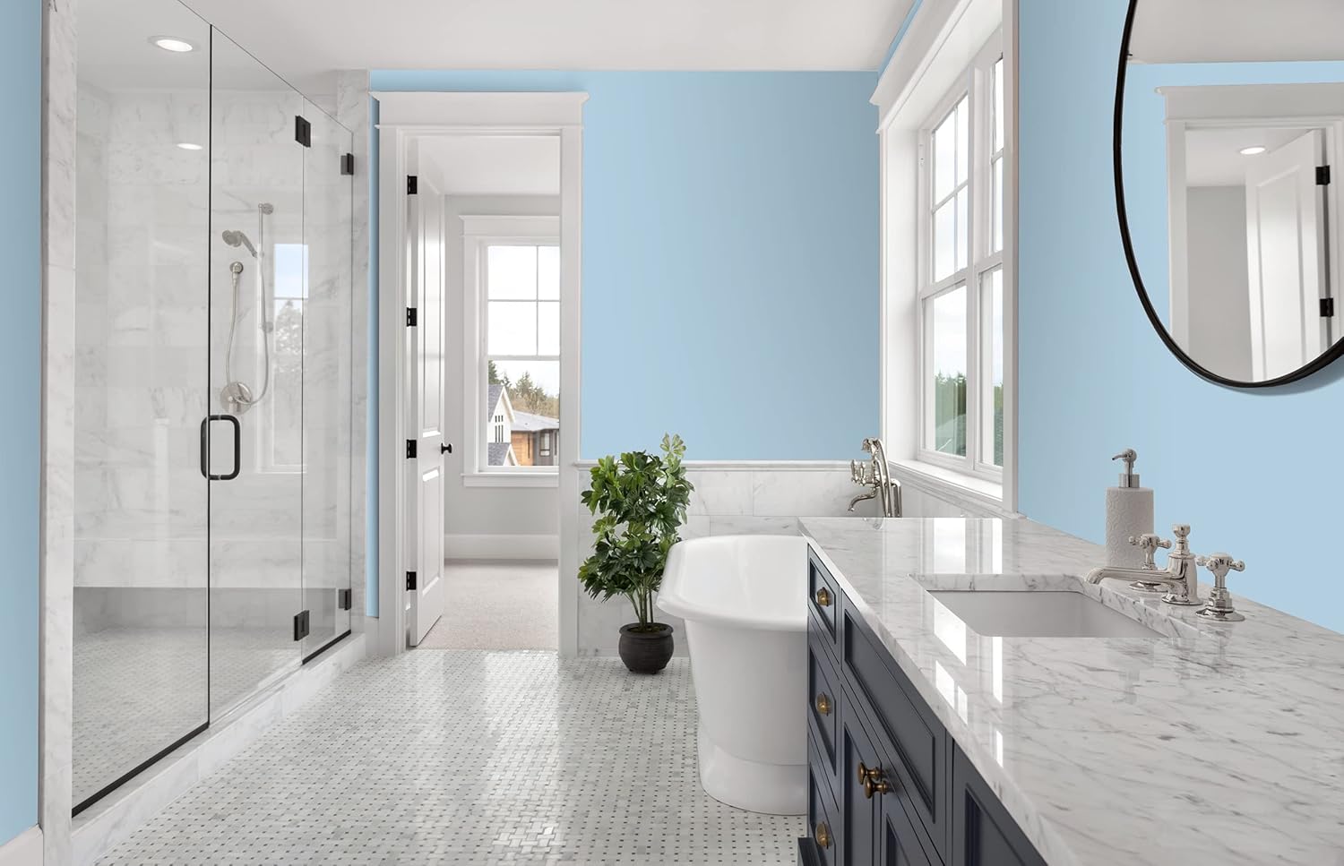 A modern bathroom with light blue premium paint on the walls, a glass shower, a freestanding bathtub, a marble countertop with dual sinks, a large round mirror, and a potted plant by the window. Natural light fills the space.