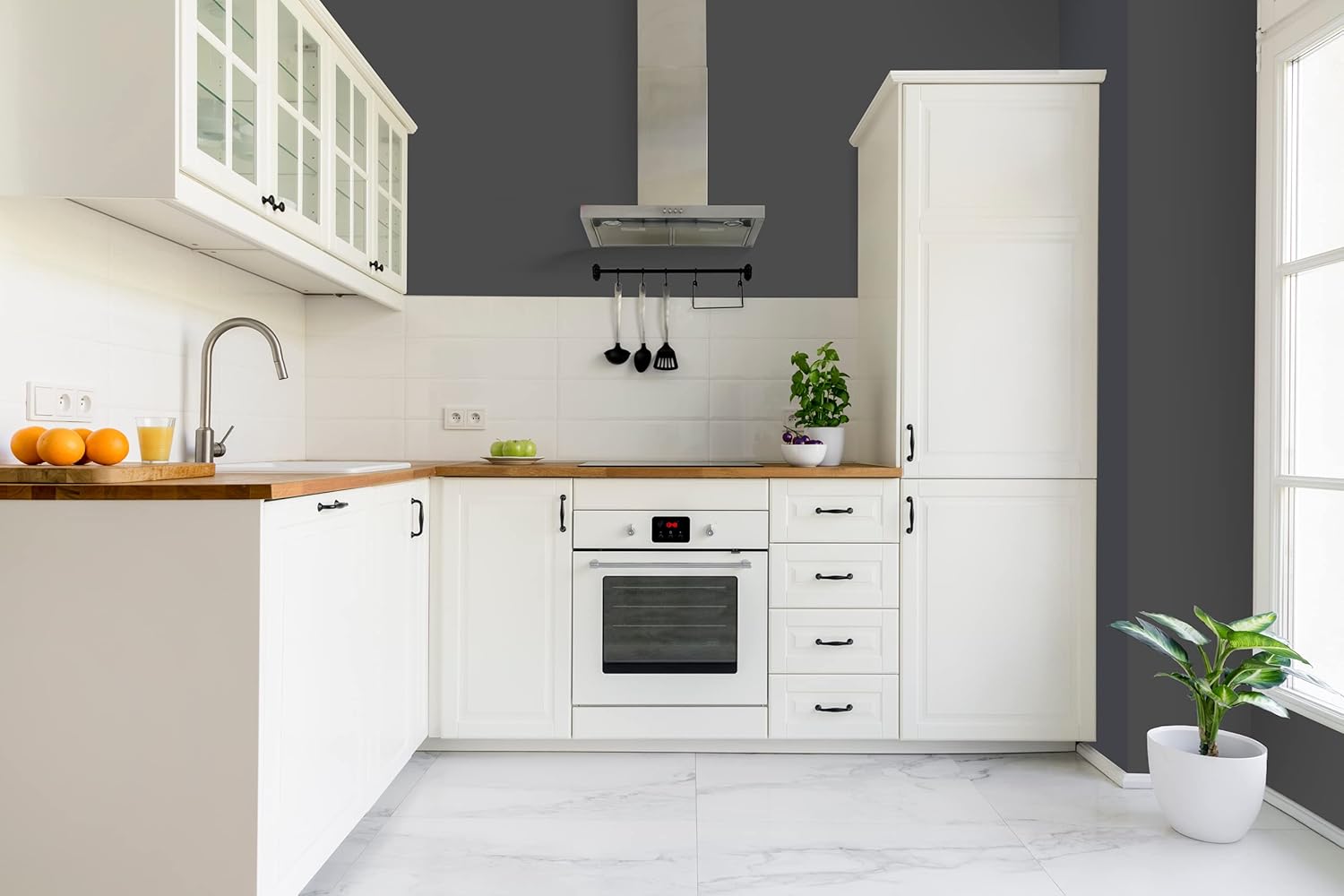 Modern kitchen with white cabinets in satin finish paint, wooden countertops, a built-in oven, stainless steel range hood, and plants. A window to the right lets natural light onto the white tiled floor.