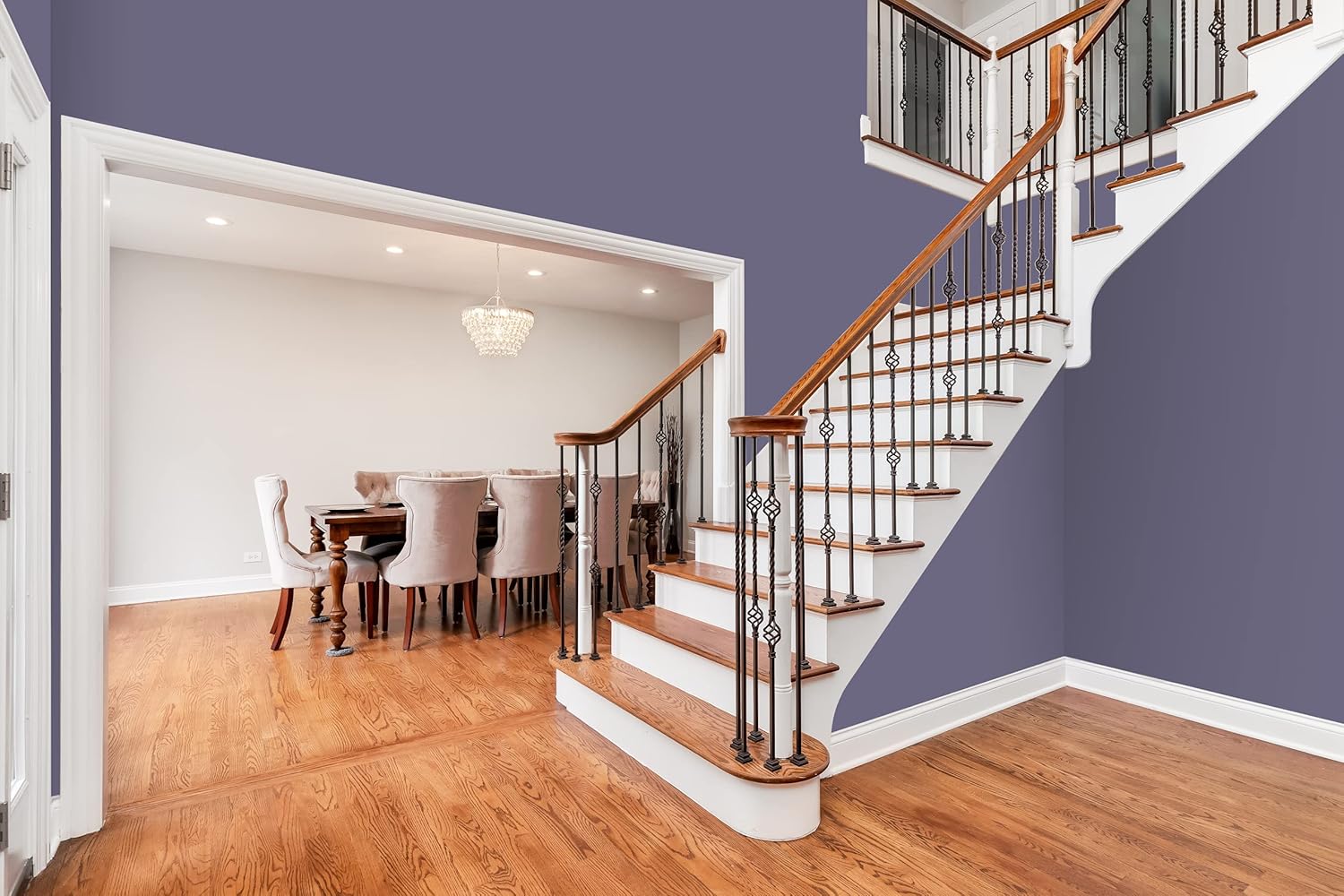 A modern home interior with wooden floors and a staircase with white risers painted in premium paint, featuring wooden handrails. The dining area has an elegant chandelier and upholstered chairs, while walls are painted a muted lavender color.