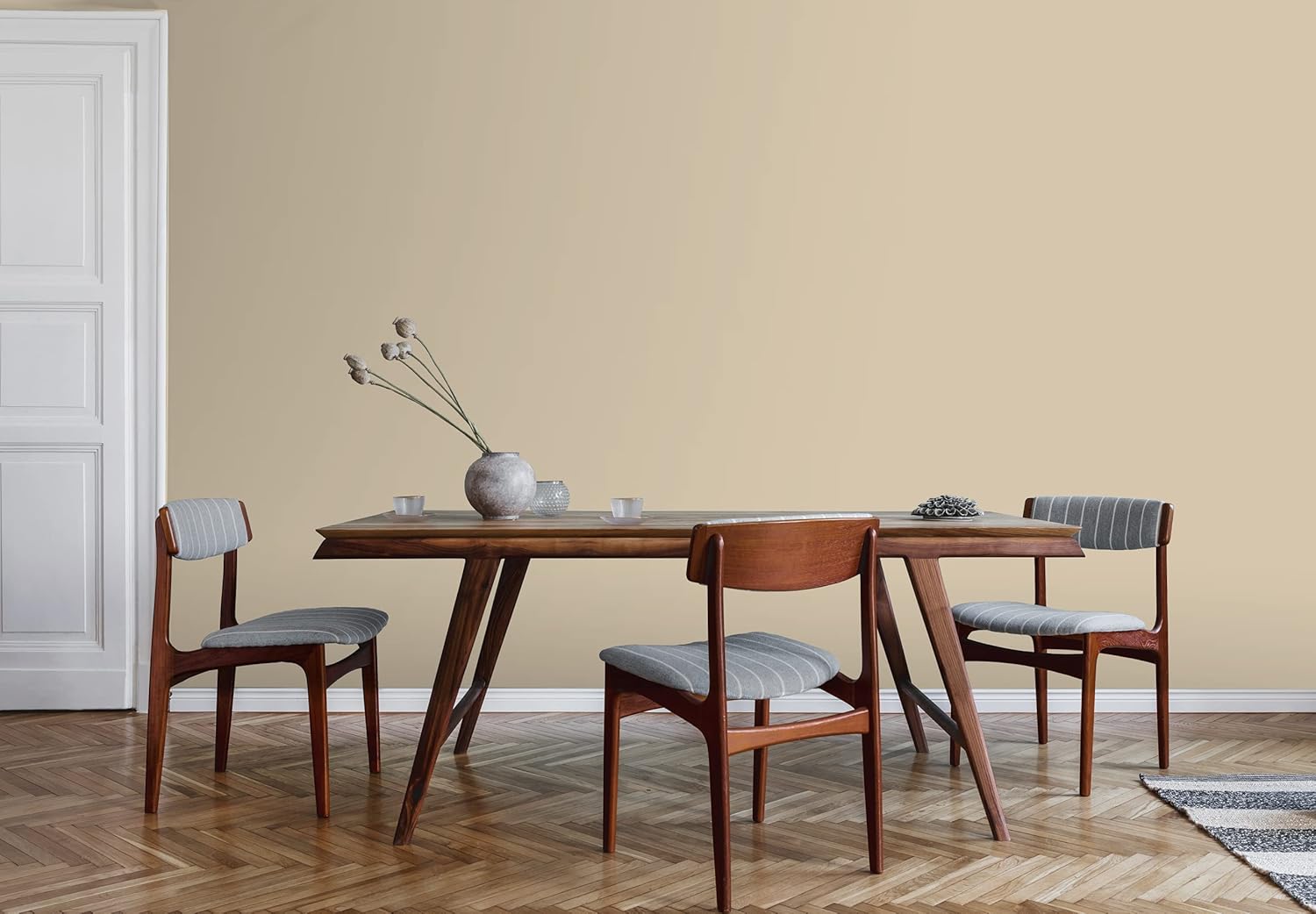 A minimalist dining room with a wooden table, three cushioned chairs, a vase with dried flowers, and a neutral-colored wall painted with premium paint. The herringbone wood floor and a portion of a rug are visible in the corner.