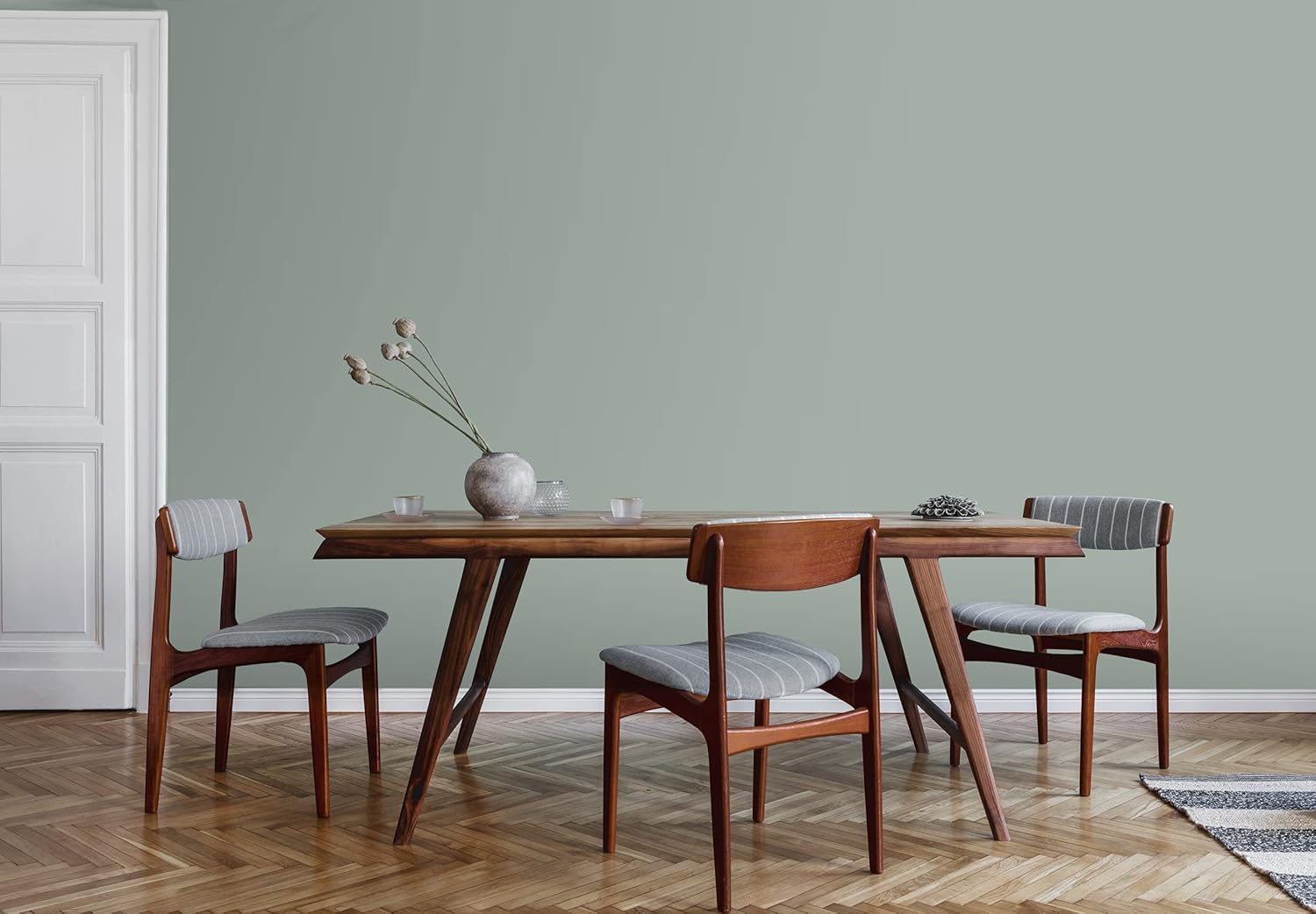 A minimalist dining room features a wooden table, three chairs with striped cushions, and a vase of dried flowers. The light green wall is coated in thick satin finish paint, perfectly complementing the chevron-patterned wooden floor.