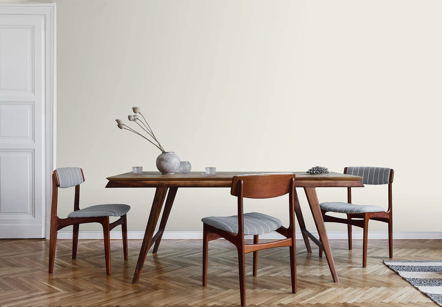 A minimalist dining room with a wooden table, three cushioned chairs, a ceramic vase, and a decorative object. The light walls feature a satin finish paint, complementing the parquet flooring and white door for an elegant, modern look.