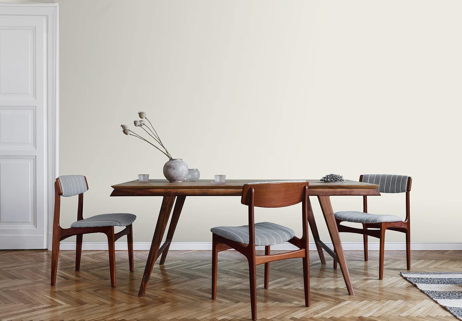 A minimalist dining room with a wooden table, three cushioned chairs, a ceramic vase, and a decorative object. The light walls feature a satin finish paint, complementing the parquet flooring and white door for an elegant, modern look.