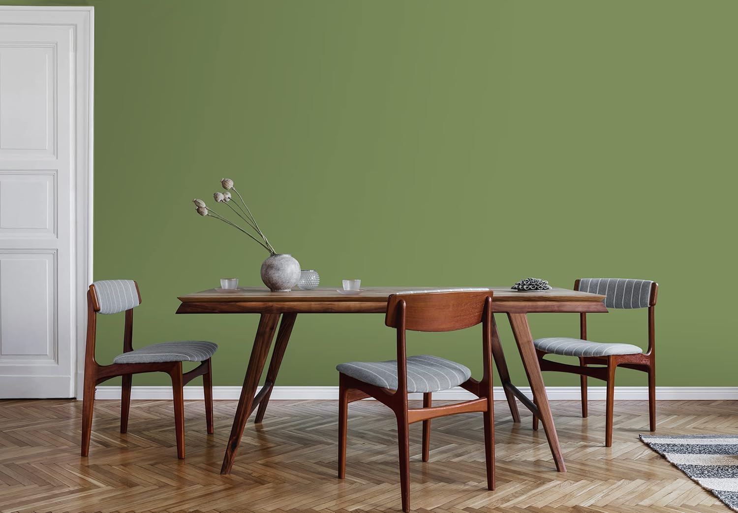 A modern dining area featuring a wooden table with a gray tablecloth surrounded by four stylish chairs. The walls are painted a calming green, and a decorative vase with dried flowers rests on the table.