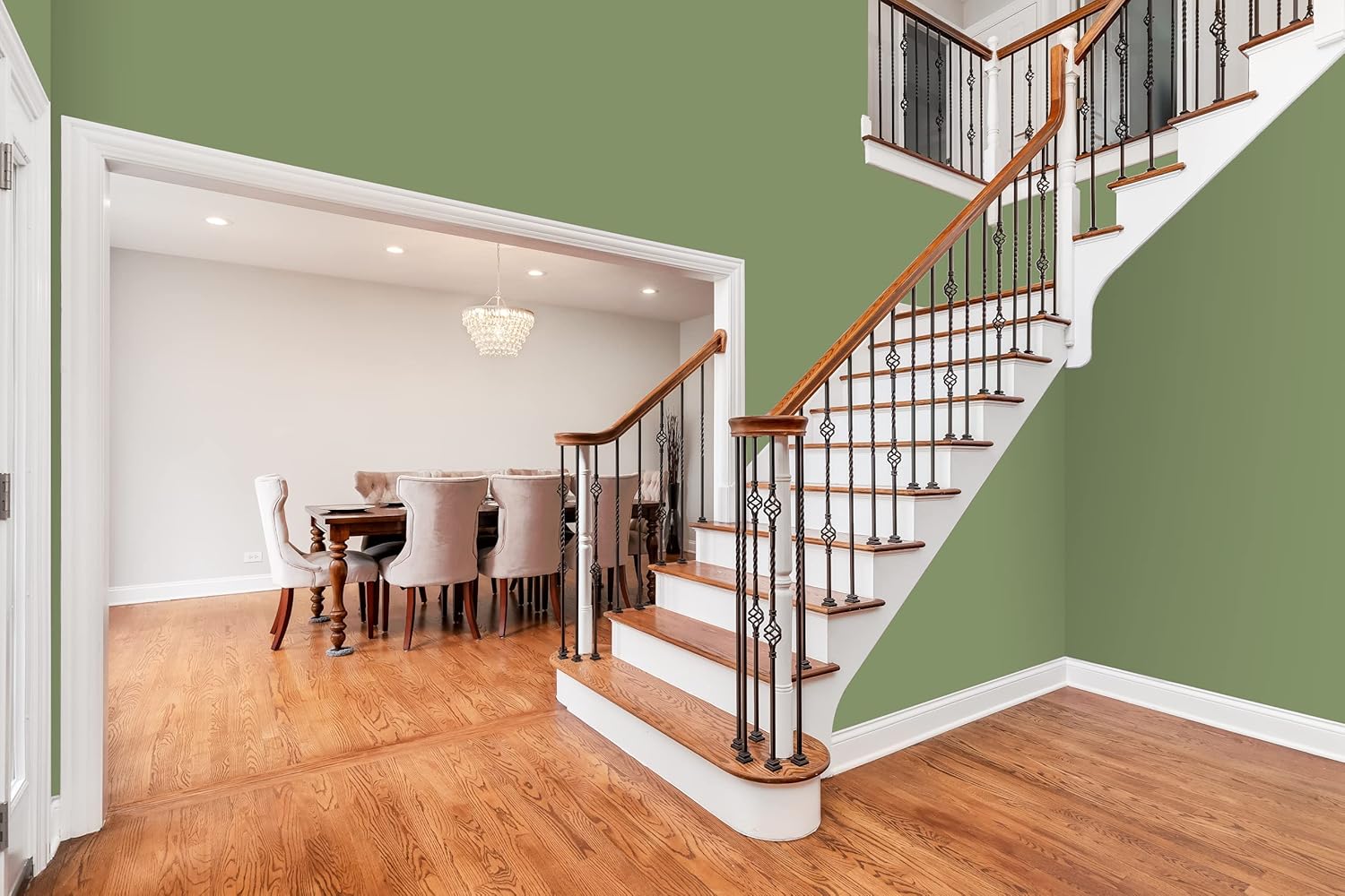 A modern entryway featuring wooden stairs with elegant black railings, leading up to a spacious dining area. The walls are painted a soft green and the floor is polished hardwood, illuminated by natural light from the dining room.