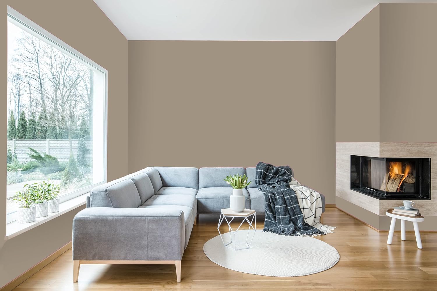 A modern living room with a large grey sectional sofa, a round white rug, potted plants, and a corner fireplace. Smooth coverage from thick satin finish paint enhances the space as natural light pours in through the large window.