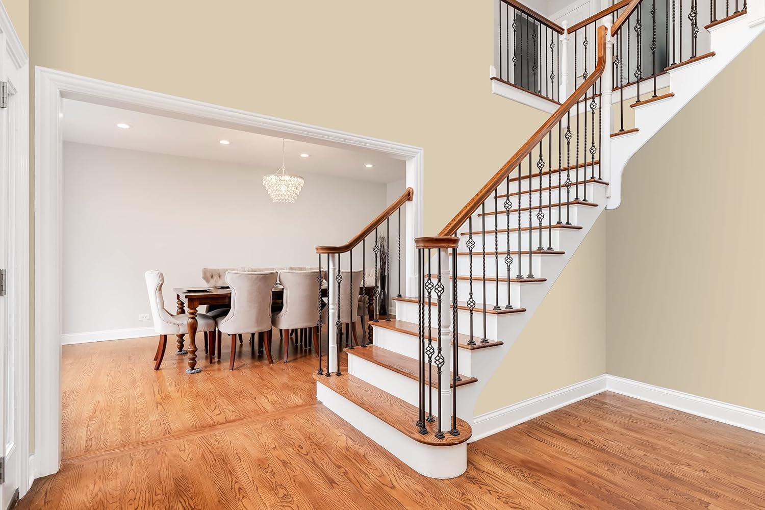 A staircase with wooden steps and black railings leads to an upper floor. Adjacent is a dining area with a wooden table, beige upholstered chairs, and a crystal chandelier. The walls feature premium interior paint in a satin finish. Floors are polished wood.