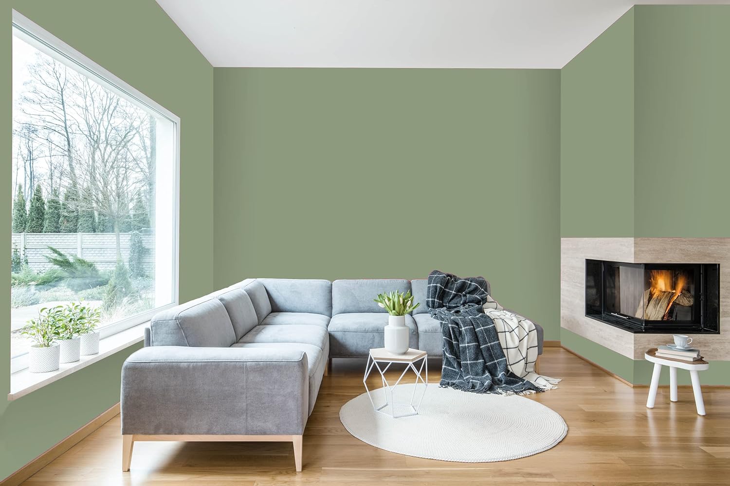A modern living room with a gray sectional sofa, a white round rug on wooden flooring, and a cozy fireplace. A vase with fresh greenery sits on a white table near a large window showcasing nature outside. The walls are painted a soft green.