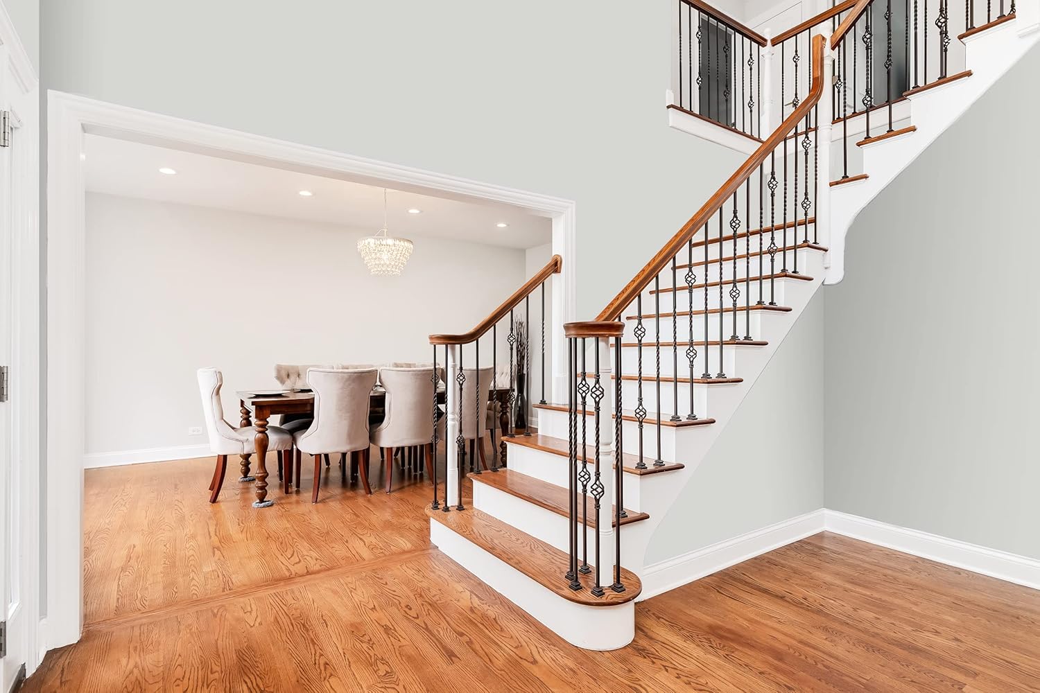 A bright foyer with hardwood flooring features a staircase with a wooden handrail and metal balusters. To the left, a dining area displays a wooden table surrounded by upholstered chairs, illuminated by a modern chandelier overhead.