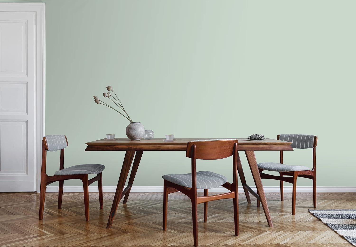A mid-century modern dining area featuring a wooden table with angled legs, surrounded by four upholstered chairs. A round vase with dried flowers sits on the table, against a soft green wall and a polished wooden floor.