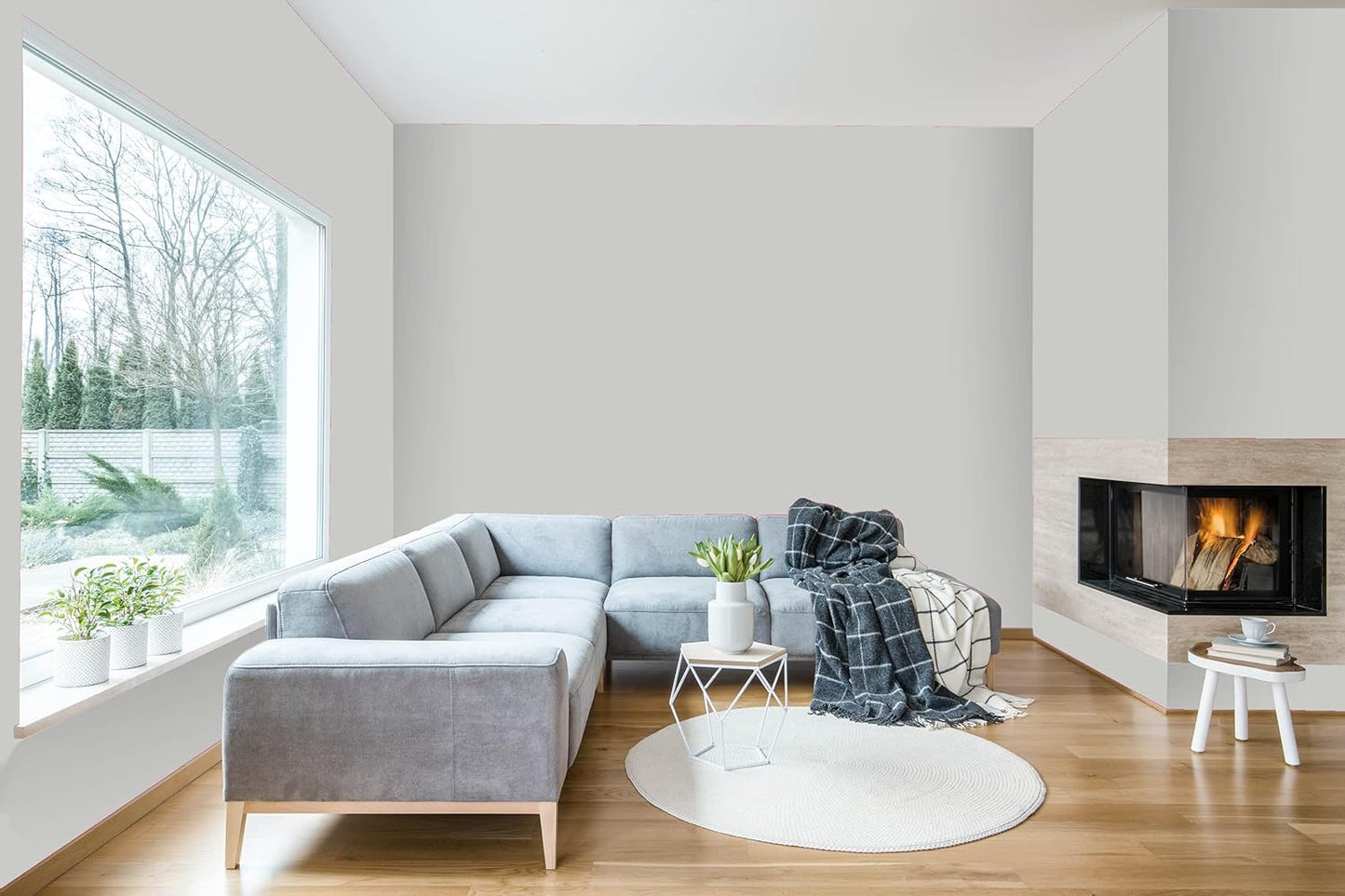 Modern living room with a large gray sectional sofa, a round white rug, cozy blanket, and a fireplace glowing warmly. The walls feature premium interior paint in a satin finish, complementing the natural light from the large window.