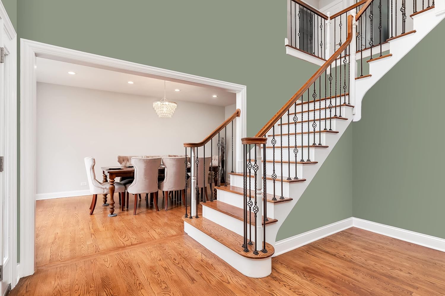 An elegant entryway featuring a wooden staircase with black iron railings and a light green wall. A dining room with a large wooden table and beige upholstered chairs is visible through an arched doorway, illuminated by a chandelier.
