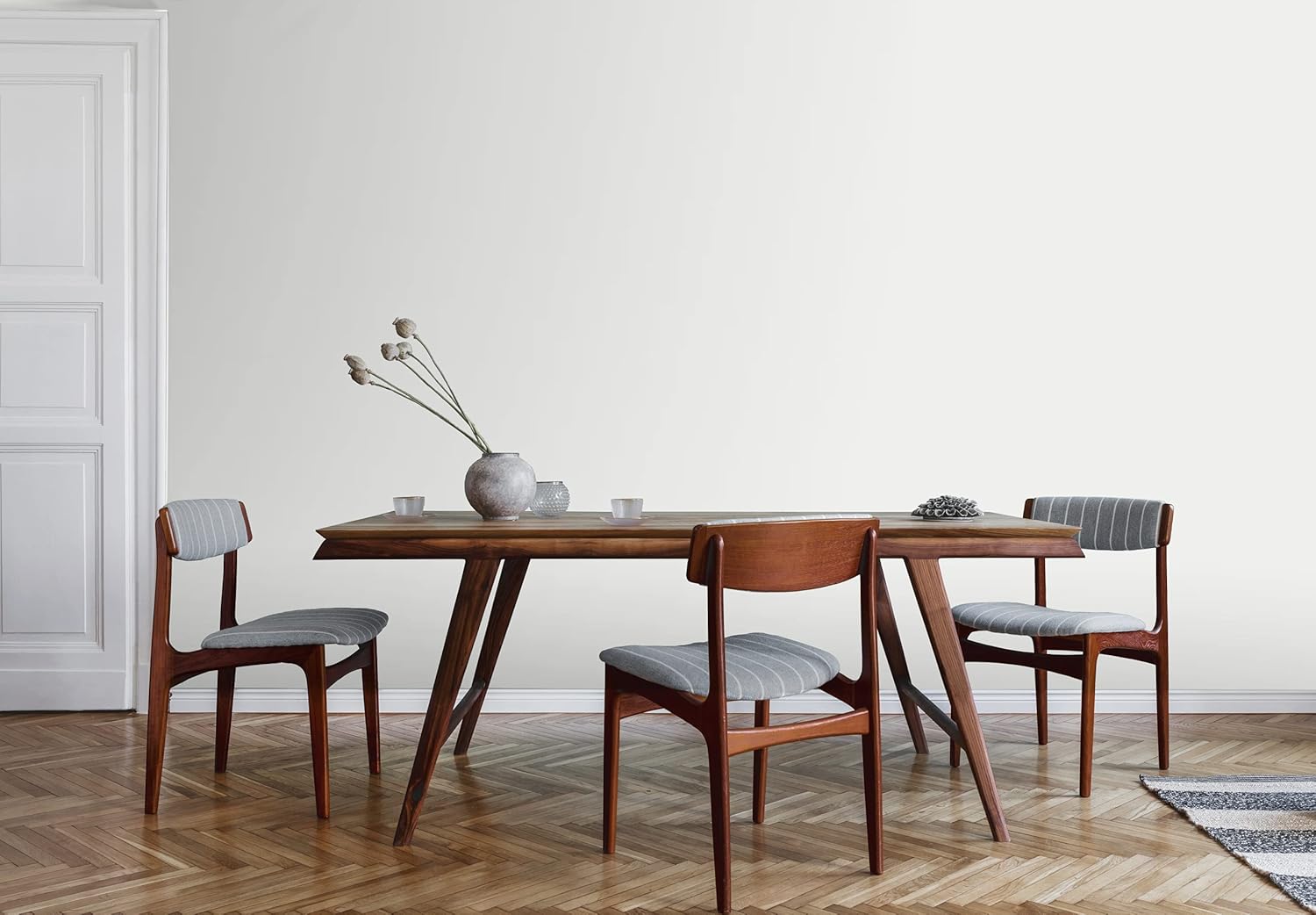 A mid-century modern dining room featuring a wooden table with angled legs and three matching chairs. A simple vase holds dried flowers on the table, complemented by minimalist decor against a light wall.
