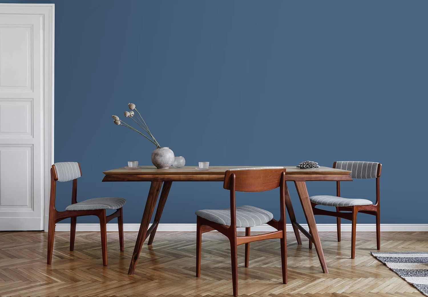 A minimalist dining room features a wooden table, three wooden chairs with gray cushions, a gray vase with dried flowers, and a blue wall coated in thick satin finish paint. The herringbone-patterned wood floor adds warmth to the serene space.
