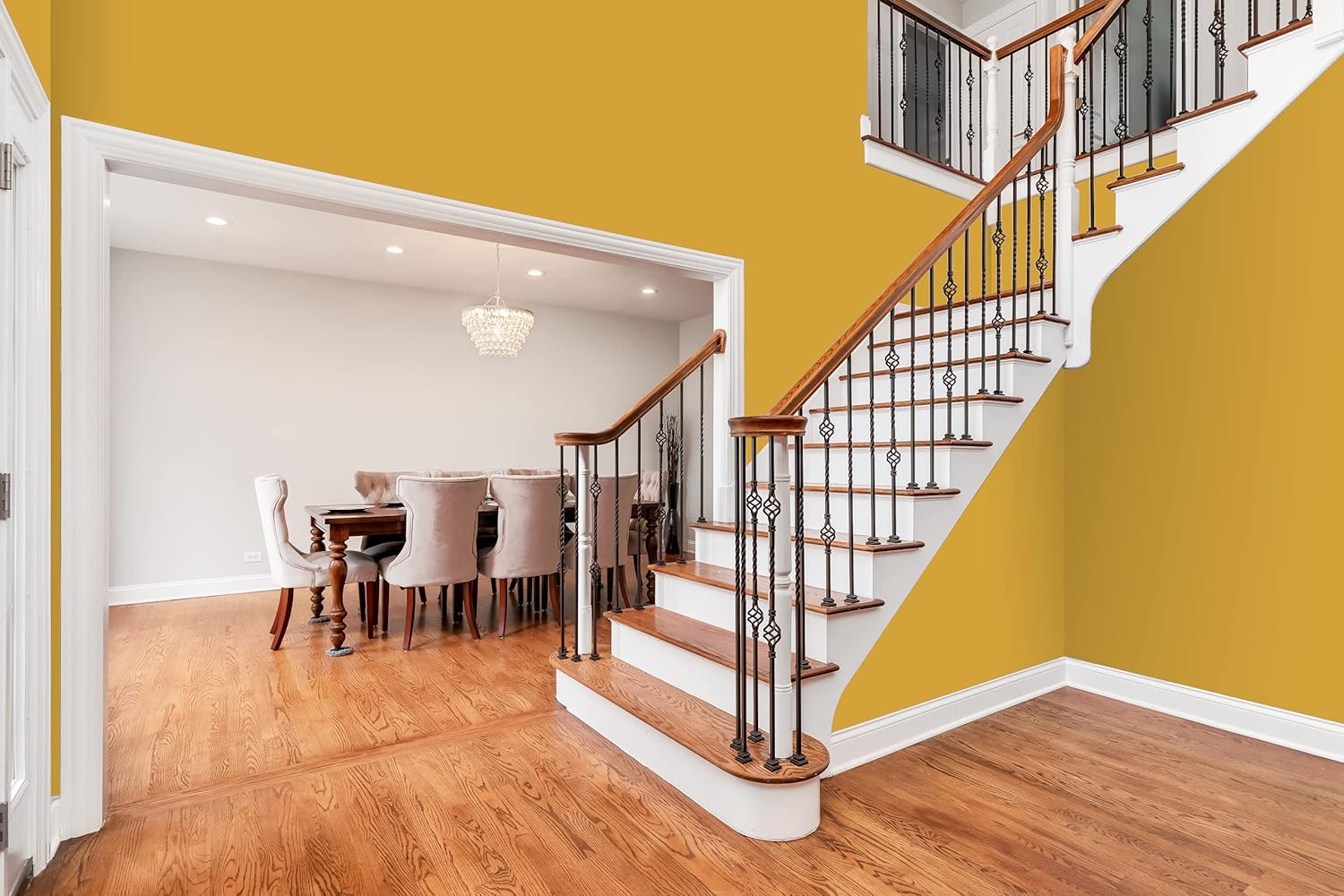 A modern home interior featuring a wooden staircase with white risers and ornate black railings, adjacent to a dining room with a chandelier and beige upholstered chairs. The walls boast a thick satin finish paint for pro-level results in mustard yellow and white.