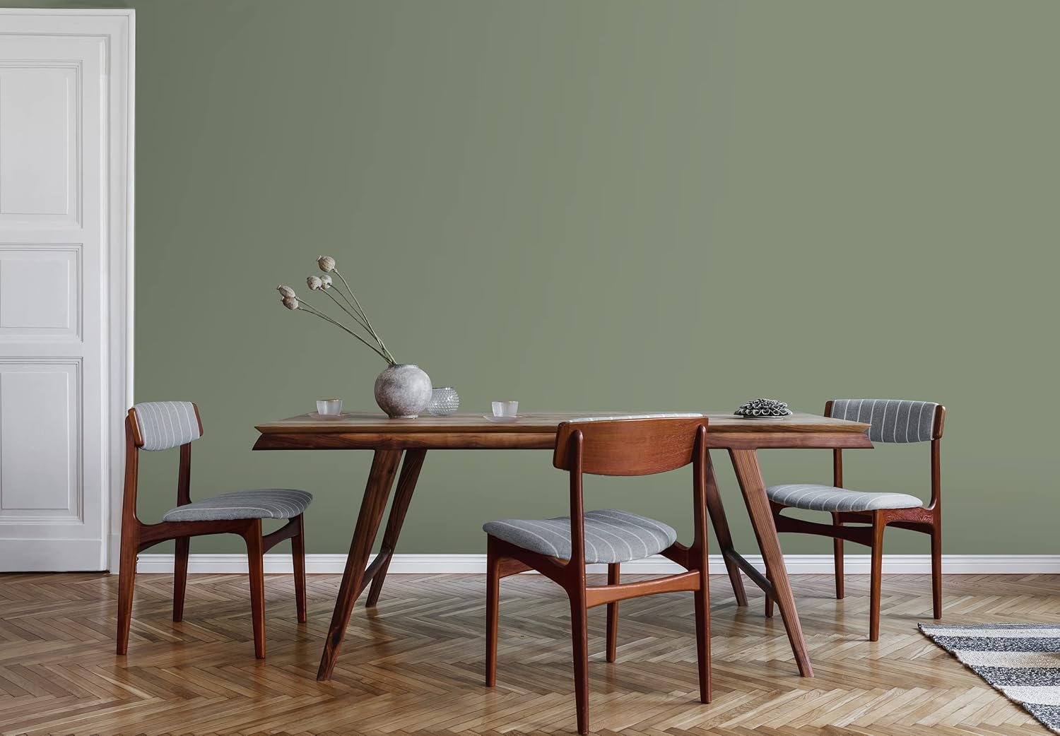 A modern dining area featuring a wooden table surrounded by four chairs, two with patterned fabric. A vase with dried flowers sits on the table, against a muted green wall, with herringbone patterned flooring and a small rug.