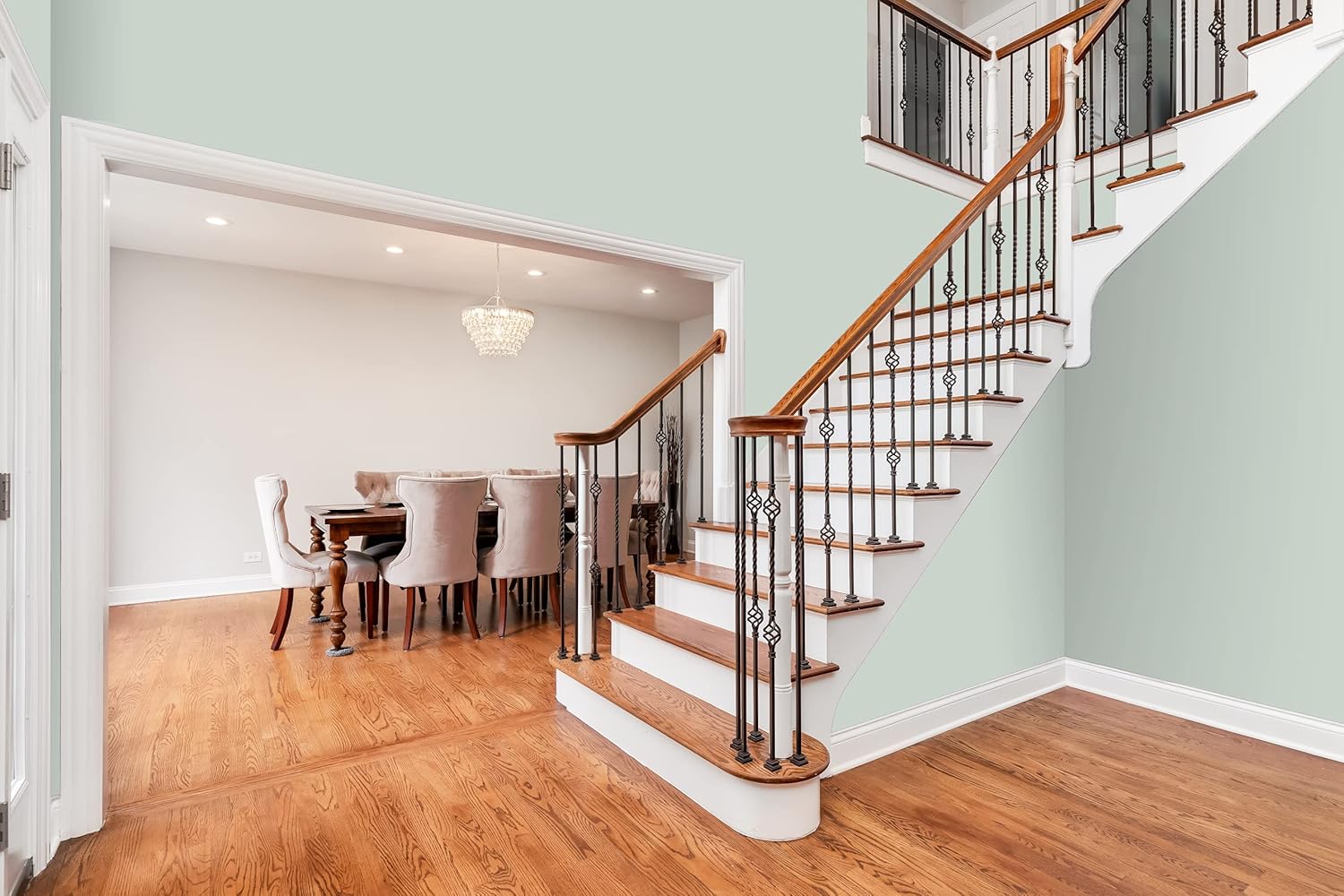 A staircase with wooden steps and a decorative railing leads to an upper floor. Adjacent is a dining area featuring bold colors, upholstered chairs, and a chandelier—all set against polished hardwood floors and light-colored premium paint walls.