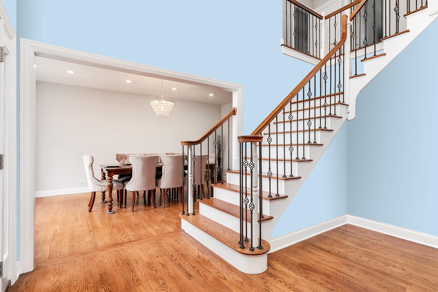 A modern, bright foyer features a wooden staircase with white risers finished in premium paint and black iron railings. Adjacent is a dining room with a table, upholstered chairs, a chandelier, and hardwood floors throughout.