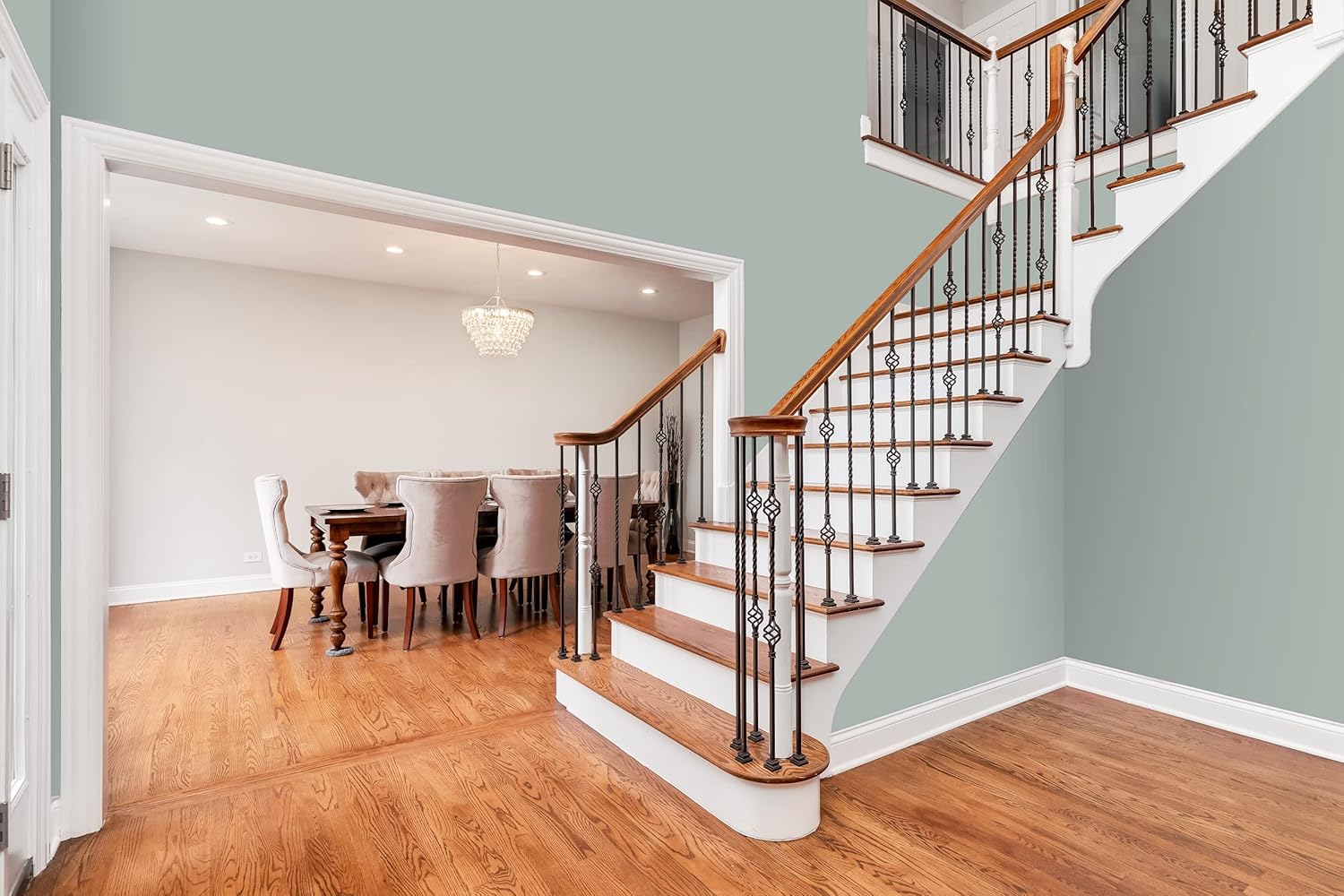 A bright, modern interior features a wooden staircase with white risers in thick satin finish paint and decorative black railings, leading up from an open area with hardwood floors to a dining room with a chandelier and upholstered chairs.