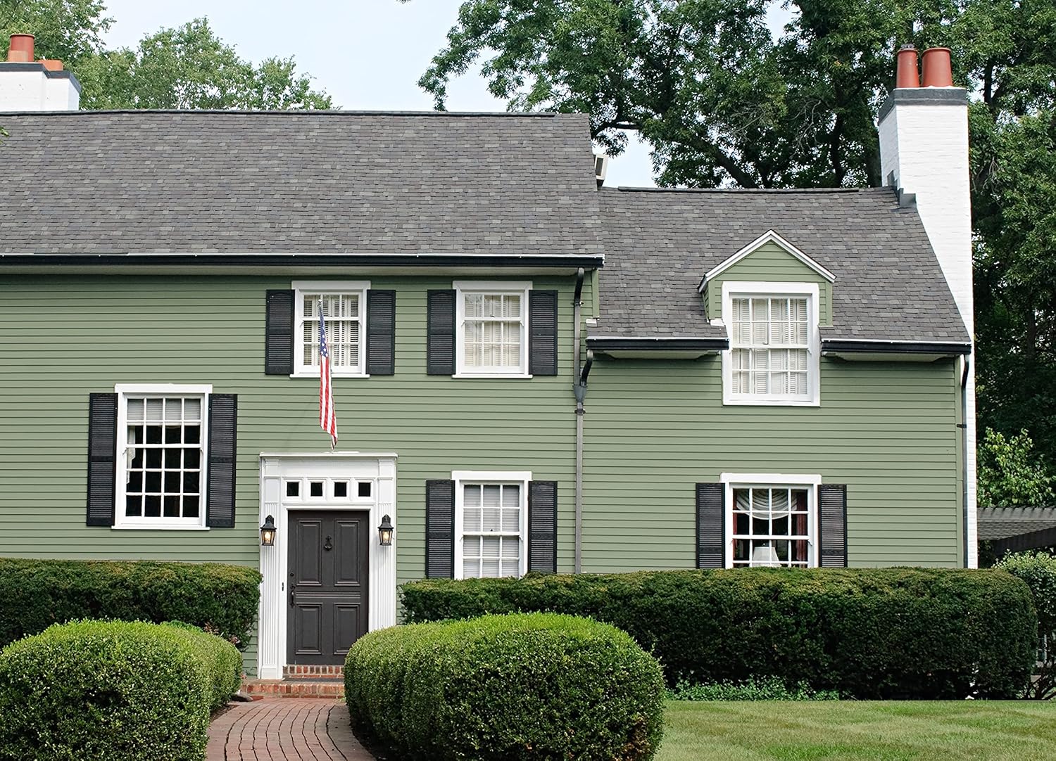 A charming green two-story house with black shutters and a white front door, framed by neatly trimmed hedges. An American flag hangs next to the entrance, adding a patriotic touch. The well-manicured lawn complements the inviting façade.