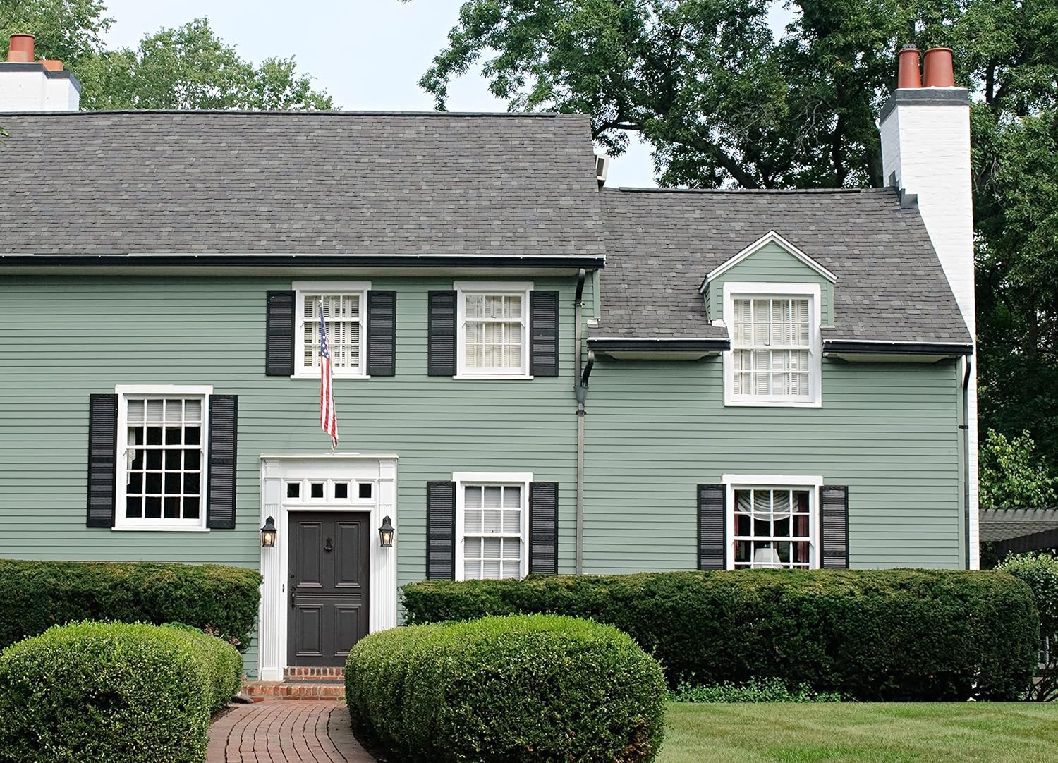 A two-story house with sage green siding coated in thick satin finish paint, white trim, black shutters, and two brick chimneys. An American flag hangs by the front door, and manicured hedges and a brick path line the front lawn.