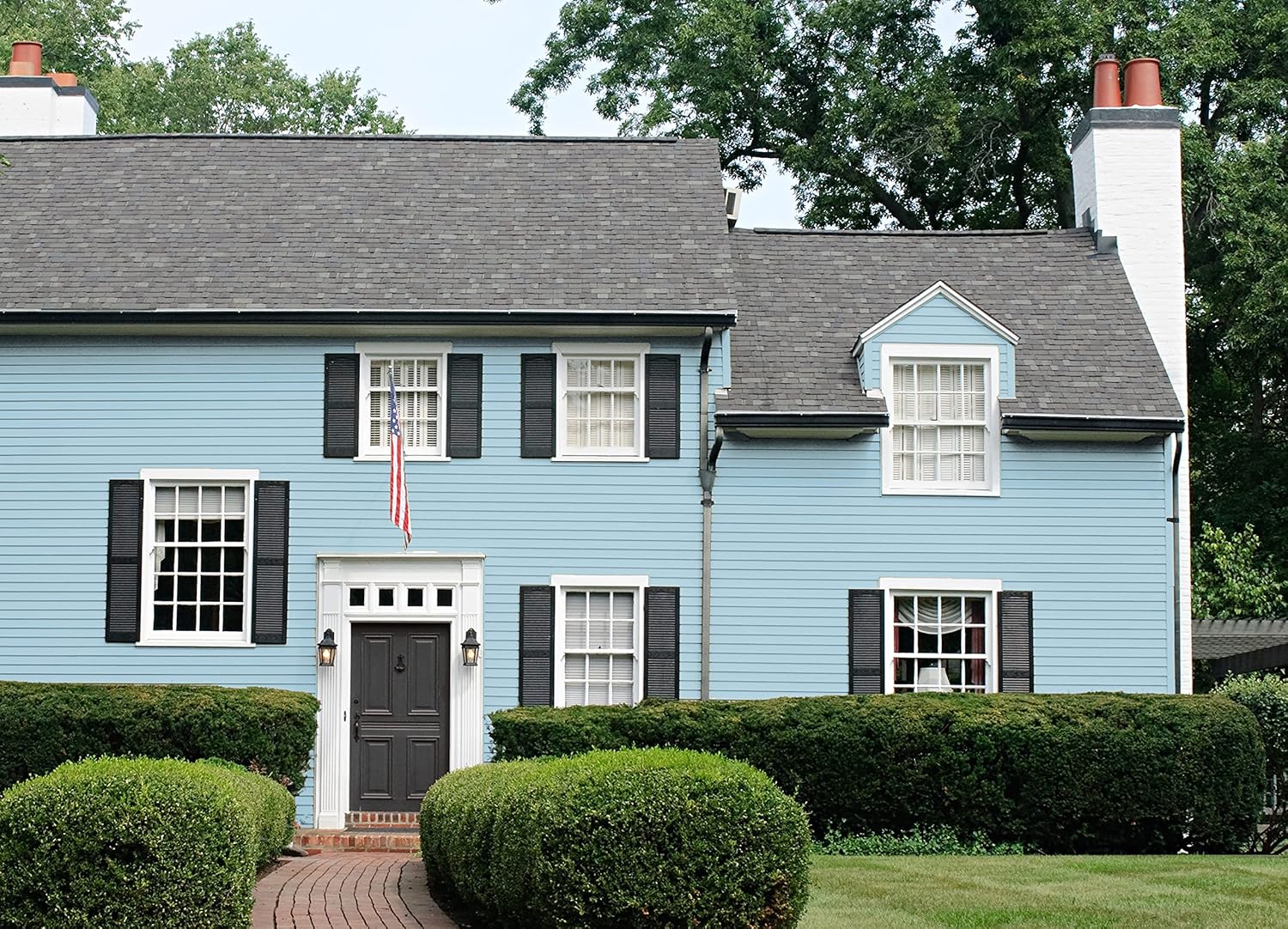 A two-story blue house with black shutters, white trim, and a dark front door painted in premium paint. An American flag hangs by the entrance. The house is surrounded by green bushes and a neatly manicured lawn.