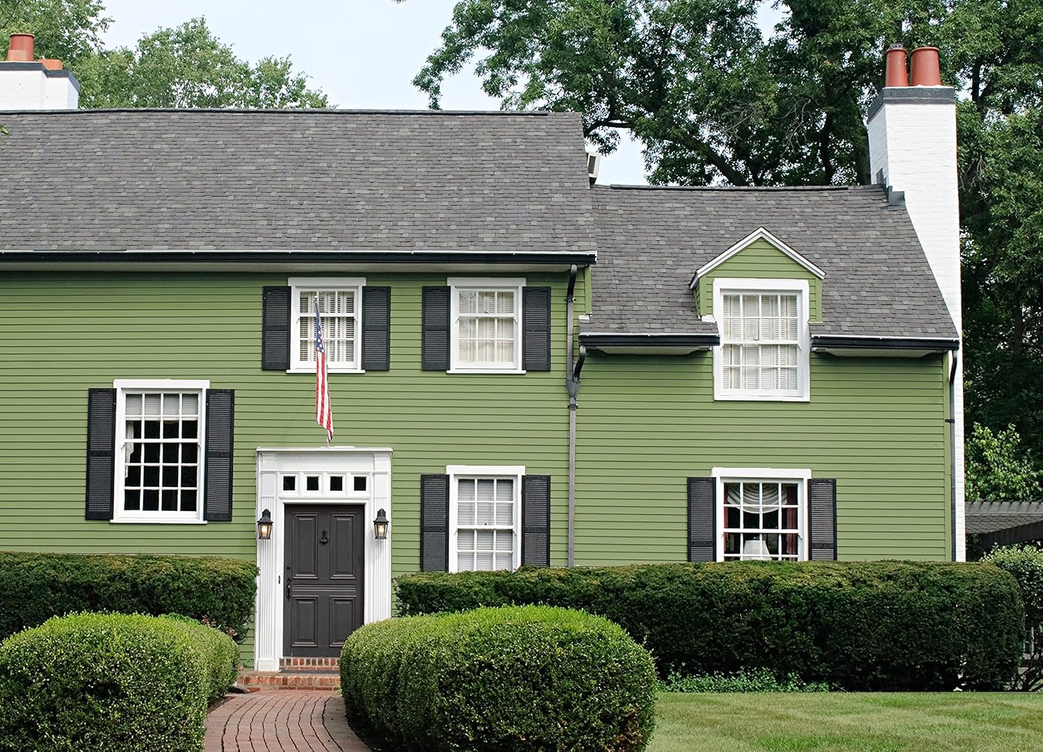 A charming two-story house with green siding and black shutters, featuring a dark front door framed by white trim. A flag hangs from the front, while neatly trimmed hedges line the walkway made of red bricks.