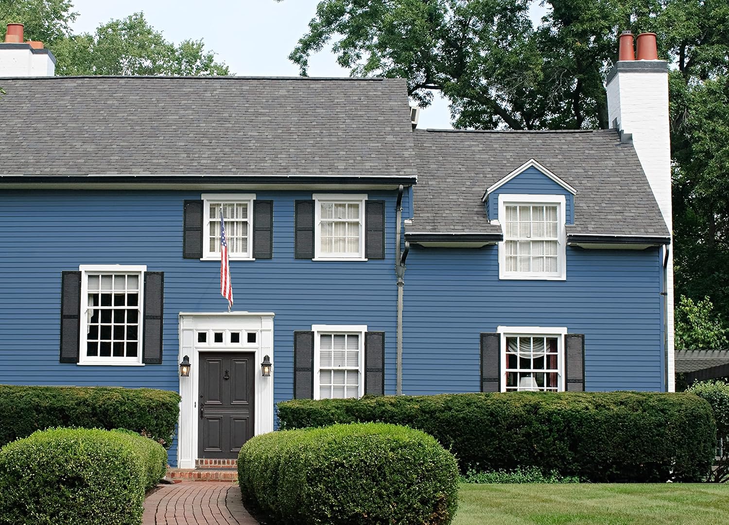 A two-story blue house with white trim, black shutters, and a gray shingled roof features a striking thick satin finish paint. An American flag hangs by the black front door, manicured hedges frame a brick path leading up to the entrance.