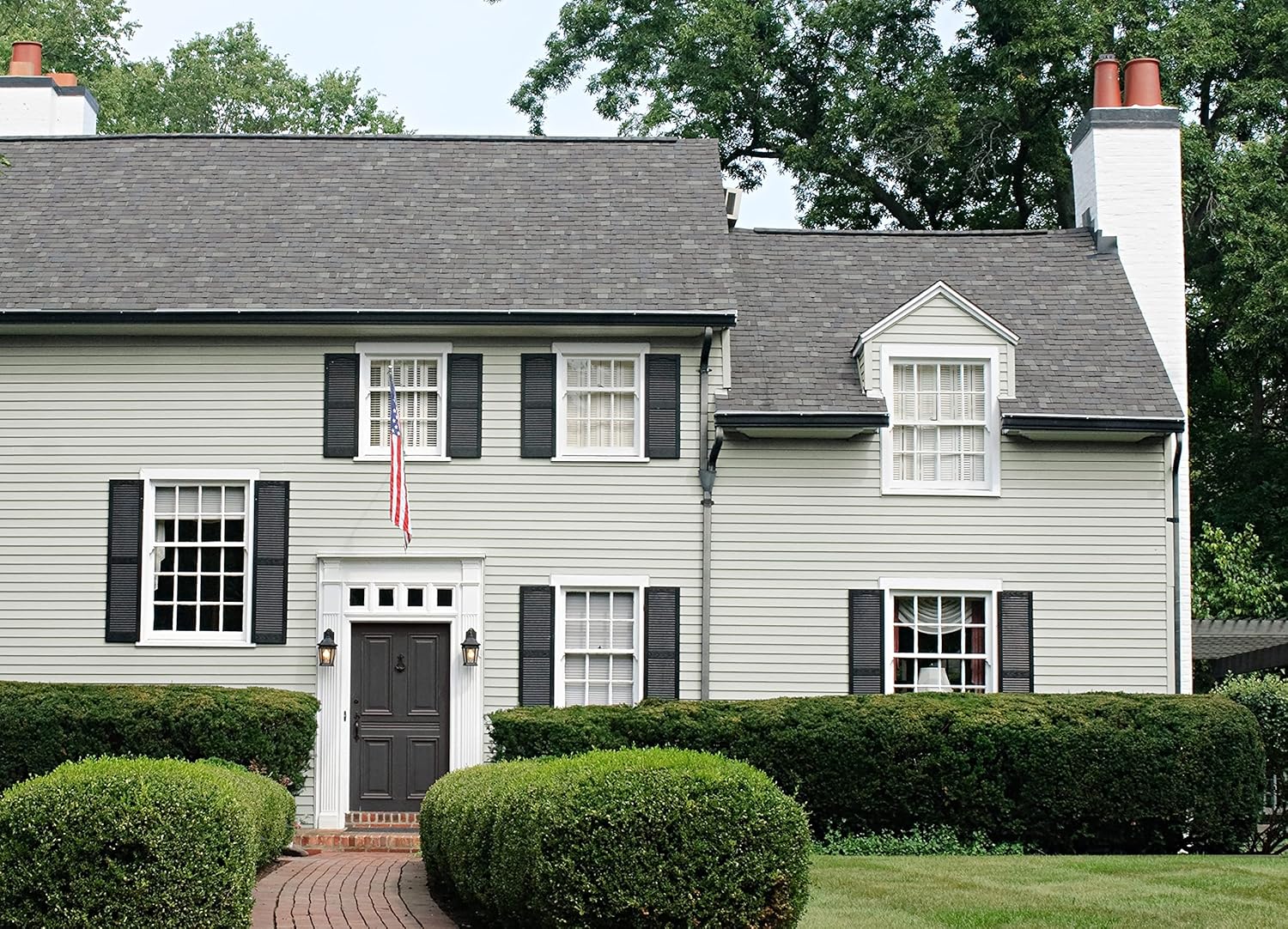 A two-story light gray house with black shutters and a front door, featuring a small American flag hanging. Lush green hedges surround the house, and a brick path leads to the entrance, while trees provide shade in the background.