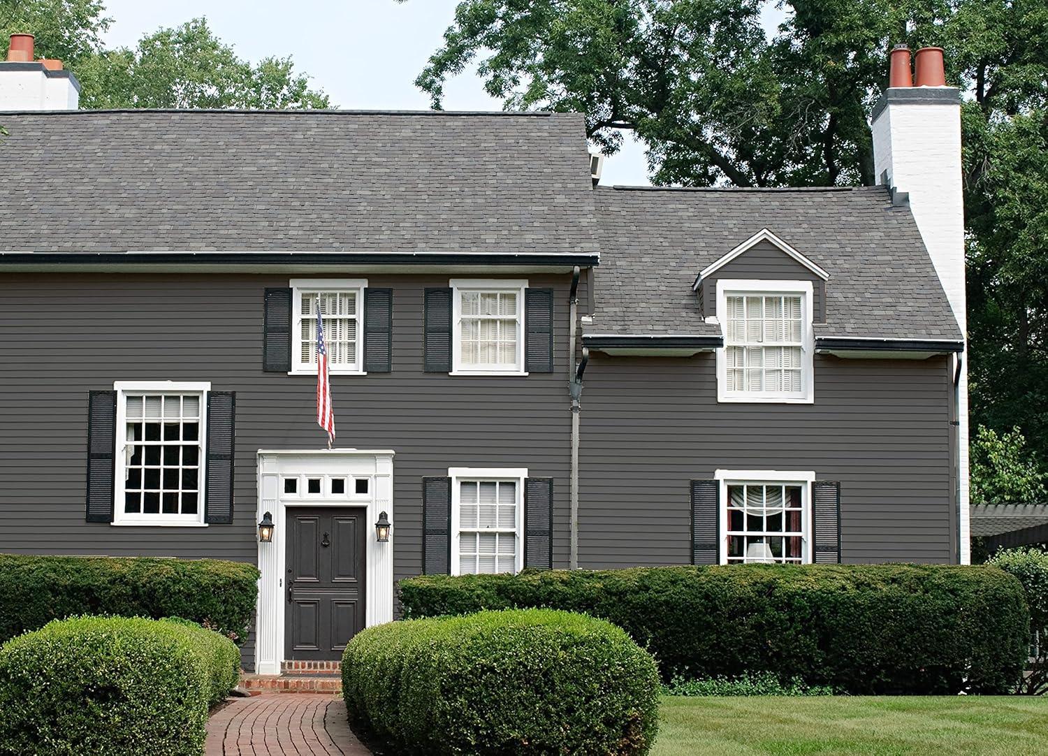 A two-story house with dark gray siding features premium interior paint, white trim, black shutters, and two chimneys. An American flag hangs above the front door, with neatly trimmed bushes and a green lawn in the foreground.