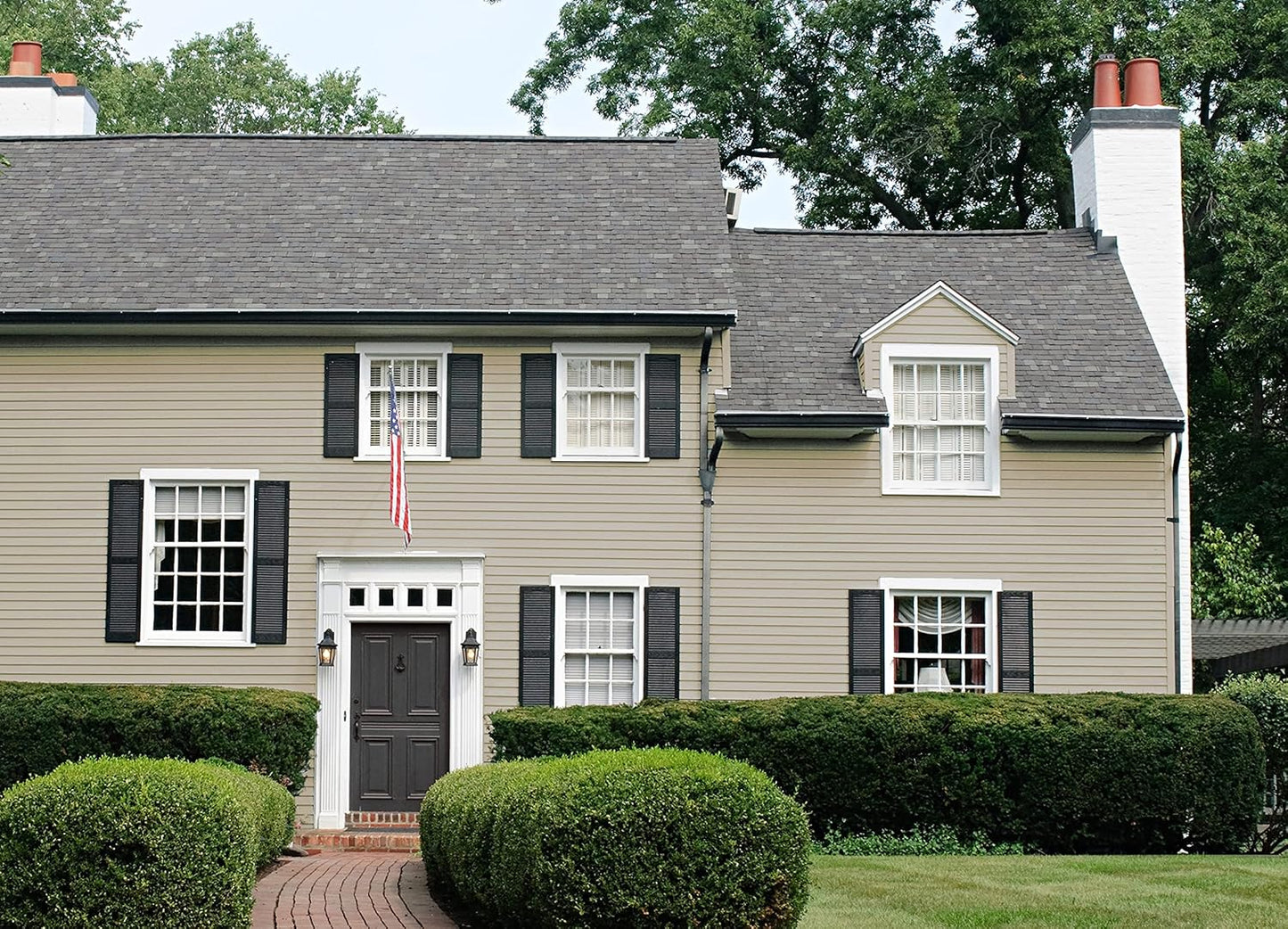 A two-story house with beige siding in a thick satin finish paint, black shutters, and a dark front door. An American flag hangs by the entrance. Well-trimmed bushes and a brick path lead to the house, with green trees in the background.