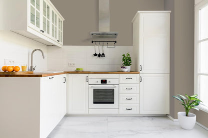Modern kitchen with white cabinets finished in thick satin finish paint, wooden countertops, built-in oven, sink, potted plants, and utensils hanging under a range hood. Sunlight streams in from a window on the right.
