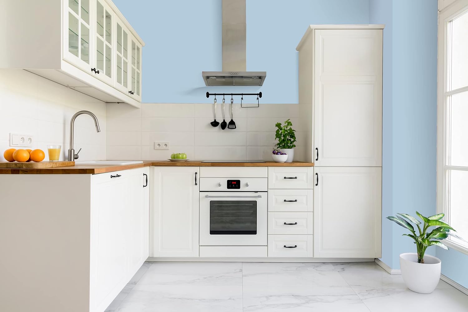 Modern kitchen with white cabinets, wood countertops, built-in oven, range hood, and sink. Light blue walls feature premium paint; a potted plant and bowl of oranges rest on the counter, with another plant near the glass door.