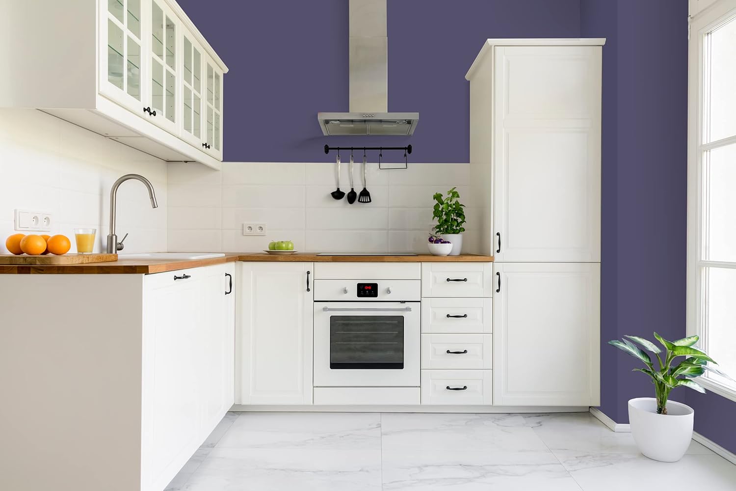 Modern kitchen with white cabinets painted in a thick satin finish paint, wood countertops, stainless steel oven and hood, utensils hanging on the wall, a potted plant on the counter, and a large window letting in natural light.