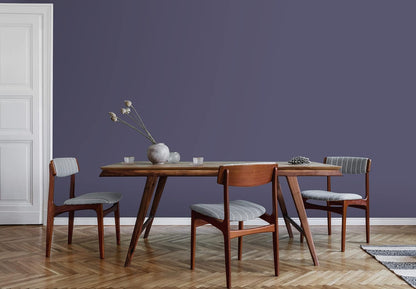 A minimalist dining room with a wooden table and three mid-century modern chairs, each with gray upholstered seats. A vase with dried flowers sits on the table. The wall features premium paint in a muted purple shade.