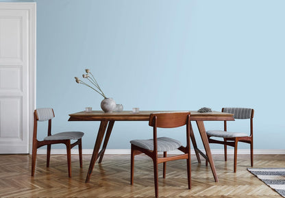 A minimalist dining room with a wooden table, three cushioned chairs, a vase with dried flowers, and a light blue wall painted in premium paint. The herringbone-patterned wood floor and white door to the left complete the serene look.