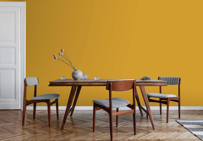 A mid-century modern dining table with four wooden chairs sits on a herringbone wood floor. A gray vase with dried flowers rests on the table, set against a mustard yellow wall painted with thick satin finish paint and a white door.