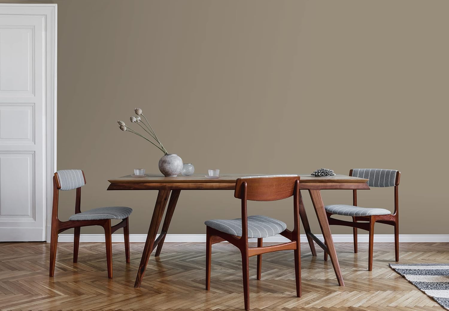 A modern dining room with a wooden table, three chairs with gray cushions, and minimalist decor on a herringbone floor stands out against a taupe wall finished in thick satin finish paint for smooth coverage. A vase of dried flowers adds charm.