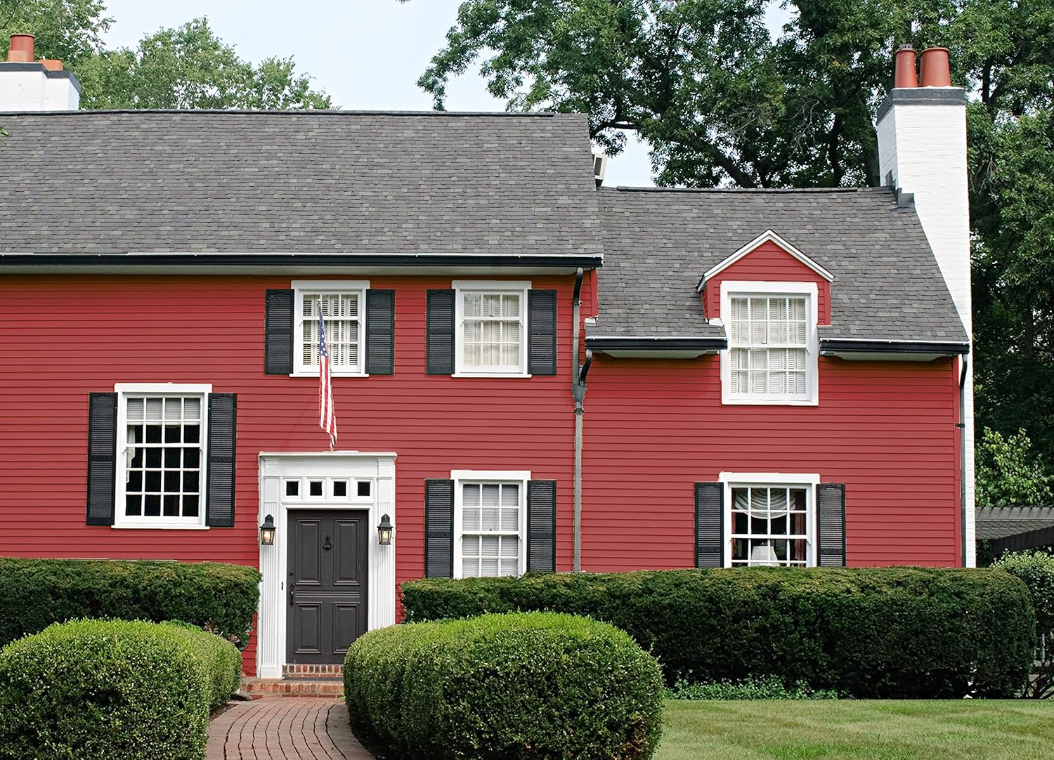 A red two-story house with black shutters, white trim, a gray shingled roof, and two chimneys. Painted with the best DIY home paint in a satin finish for lasting beauty. An American flag hangs by the black front door amid neat bushes and a green lawn.