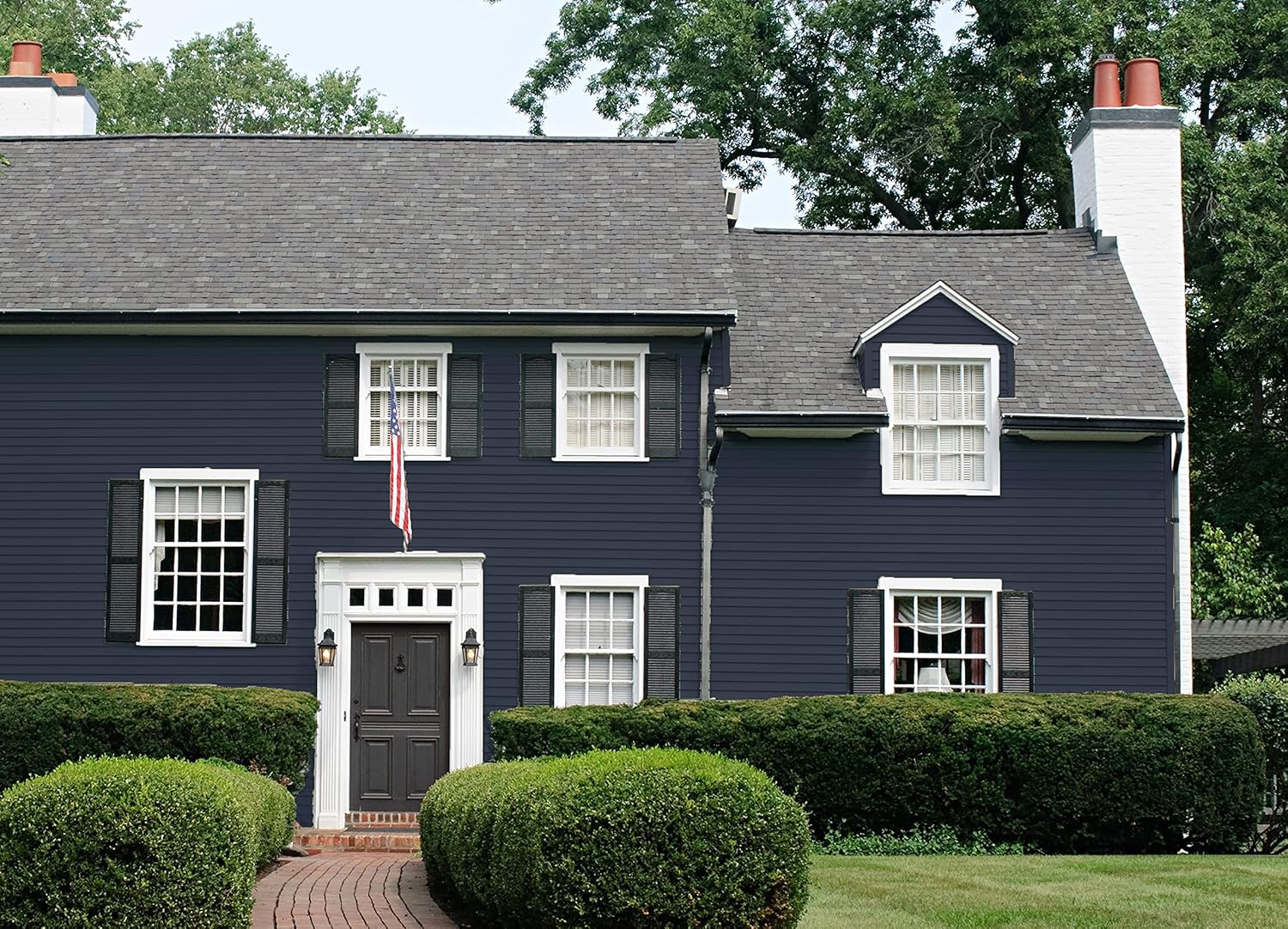 A two-story house with dark blue siding painted with the best DIY home paint, white trim, black shutters, and a gray shingled roof. A small American flag hangs by the front door, and the yard has neatly trimmed bushes and a brick walkway.